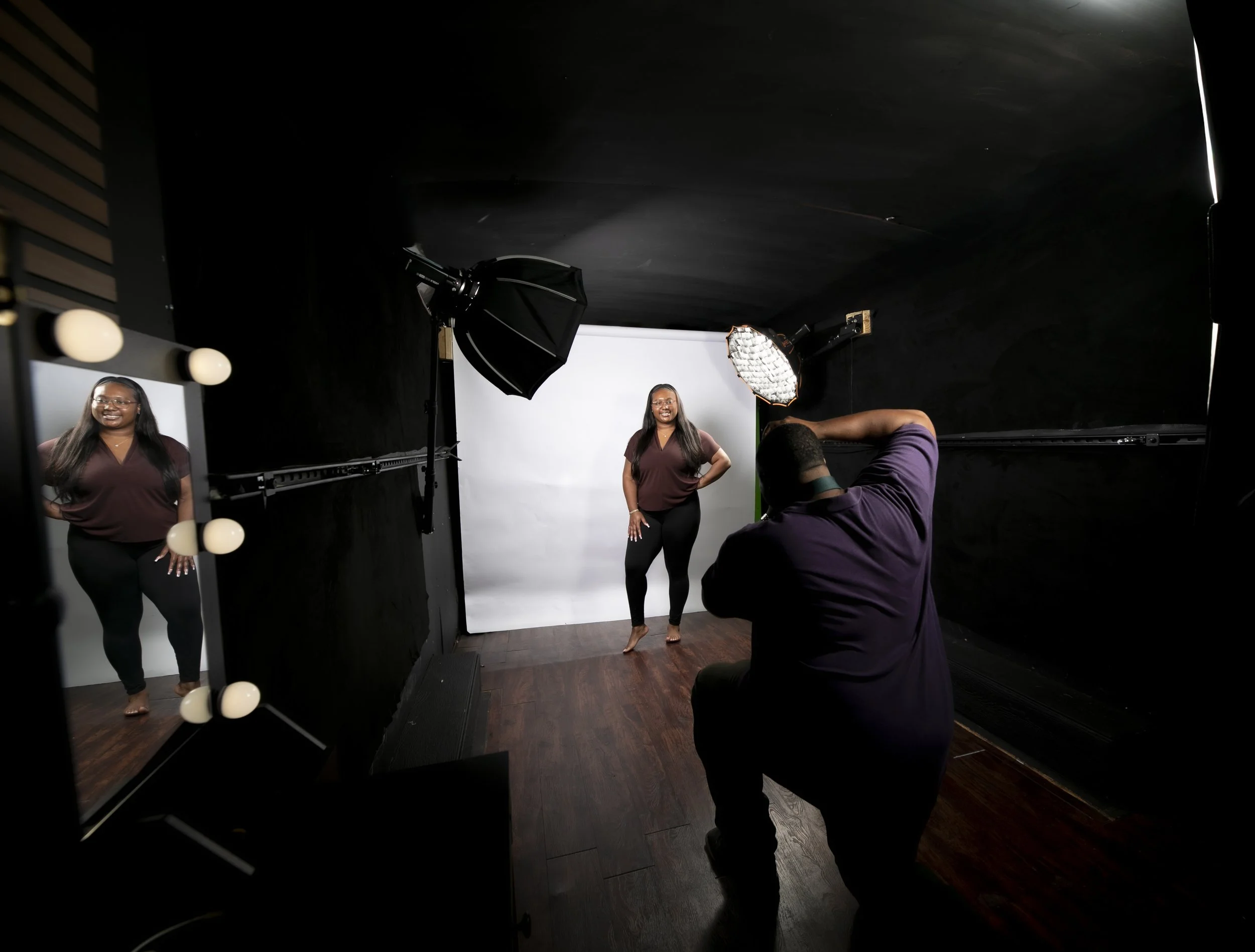 A woman standing in front of a white backdrop in a photography studio, posing for a photoshoot. A photographer is taking her picture. The studio has professional lighting equipment, including a large softbox light and an umbrella reflector.