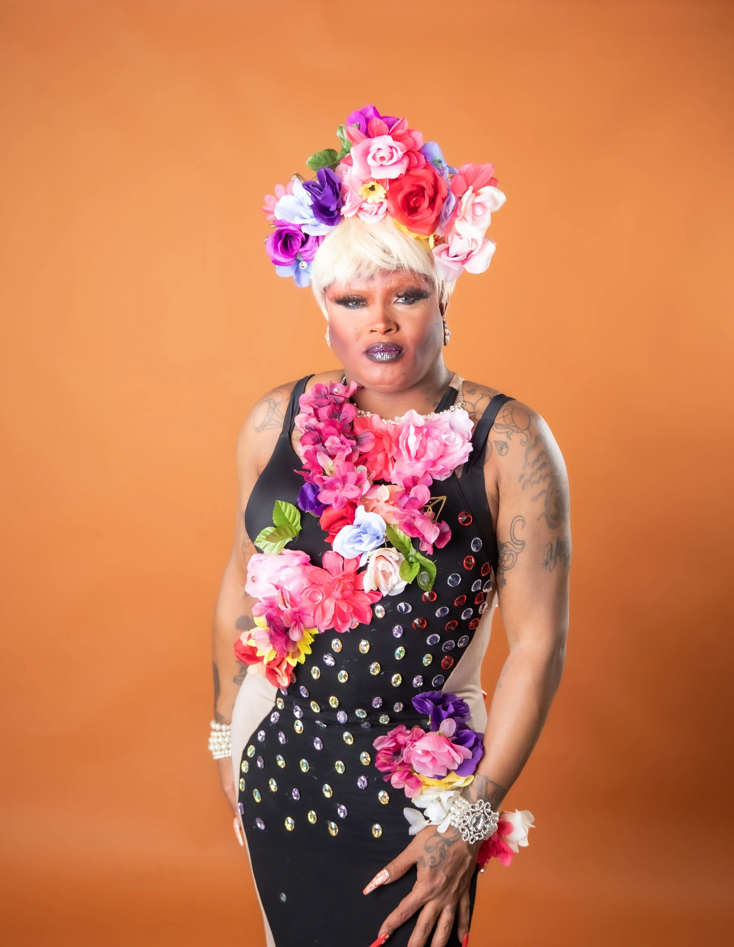 A person with light skin and platinum blonde hair styled in a short bob, wearing a large floral headdress made of pink, red, purple, white, and blue flowers. The individual is dressed in a black outfit decorated with shiny circular embellishments and a pink floral necklace. They have tattoos on their arms, sparkly makeup, and jewelry including earrings, a bracelet, and a ring. The background is a solid orange color.