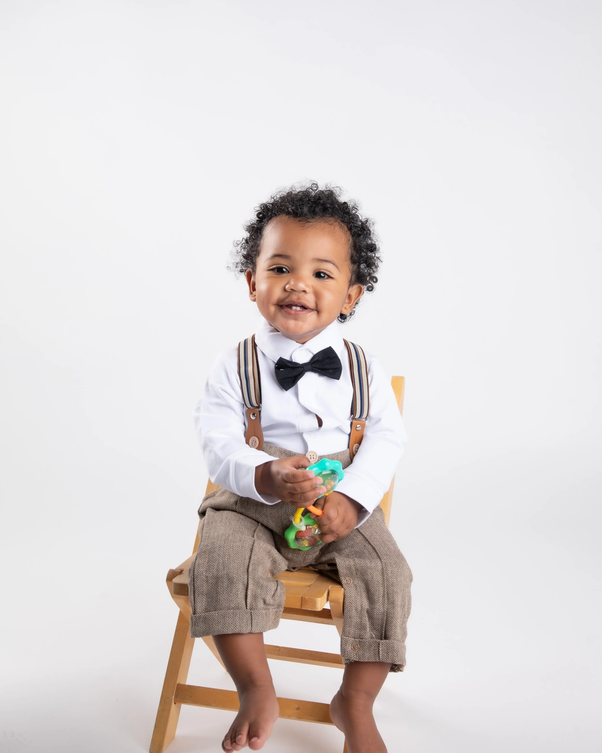 A young child with curly black hair, wearing a white shirt, black bow tie, suspenders, and brown plaid shorts, sitting on a wooden chair against a plain white background, smiling and holding a toy.