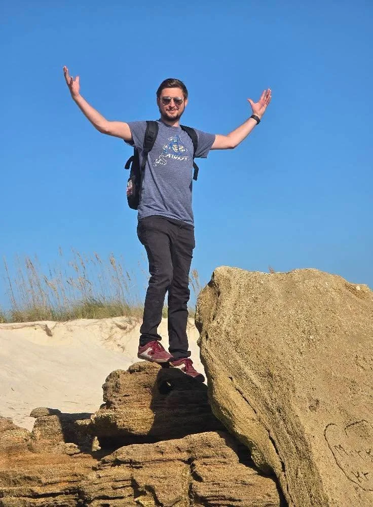 A man wearing sunglasses, a blue t-shirt, and black pants standing on a large rock with arms raised in a celebratory pose outdoors against a clear blue sky.