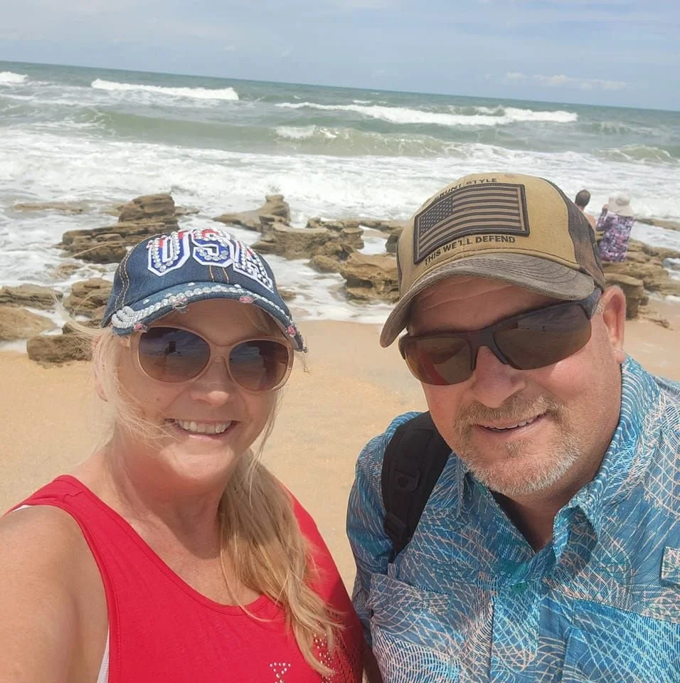 A smiling woman in a red top and sunglasses, and a smiling man in a blue patterned shirt and sunglasses, taking a selfie on a beach with rocks and ocean waves in the background.