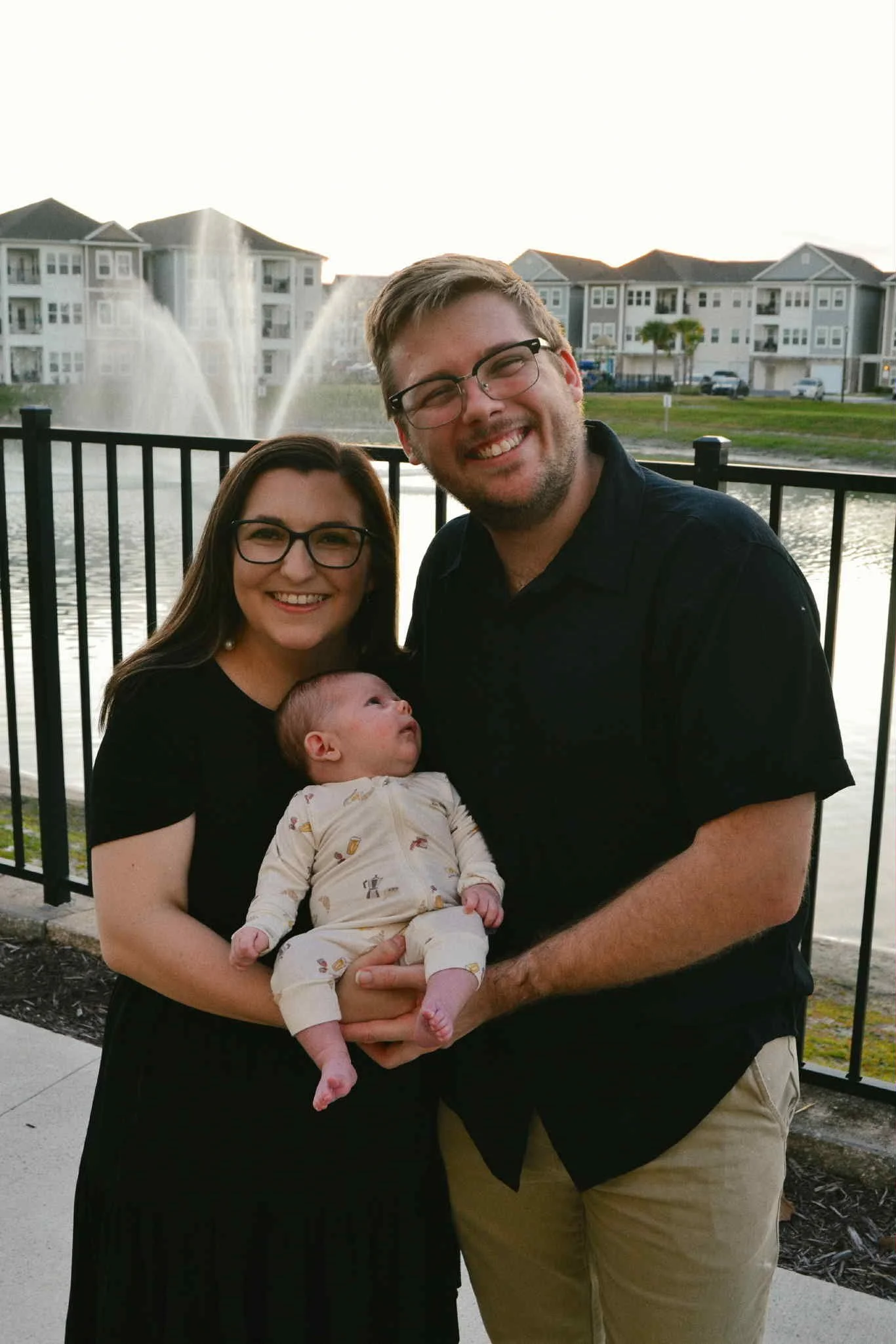 A family of three posing outdoors by a lake with a fountain and apartment buildings in the background. The woman is holding a baby, and the man is standing beside them, all smiling.