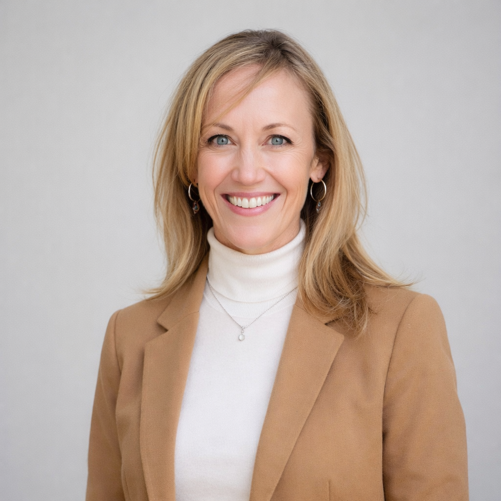 A smiling woman with blonde hair wearing a beige blazer and white turtleneck, standing against a plain light gray background.
