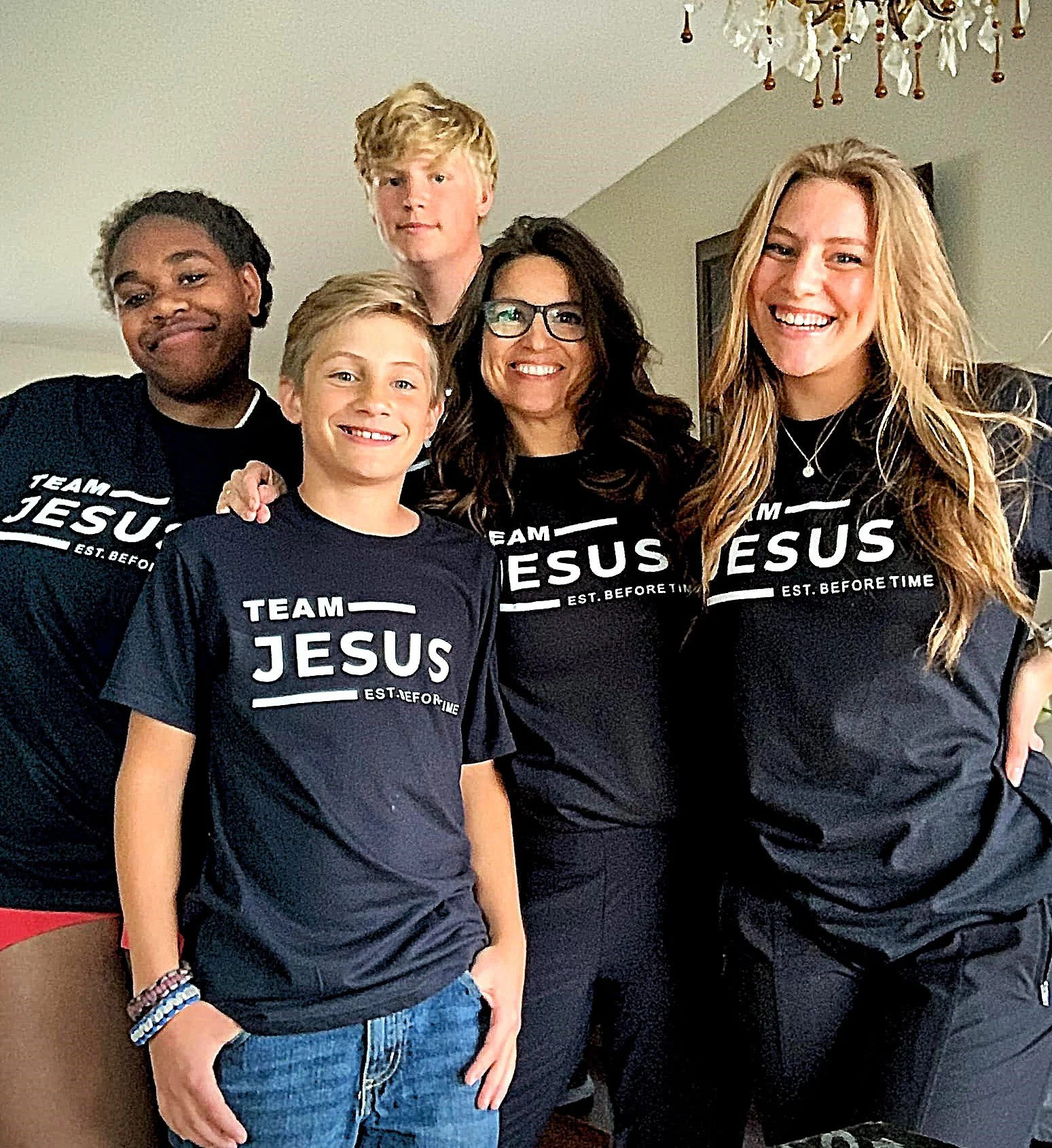 Group of five people, three females and two males, smiling and posing together indoors. Four of them are wearing matching black T-shirts that say "TEAM JESUS" and "EST. BEFORE TIME."