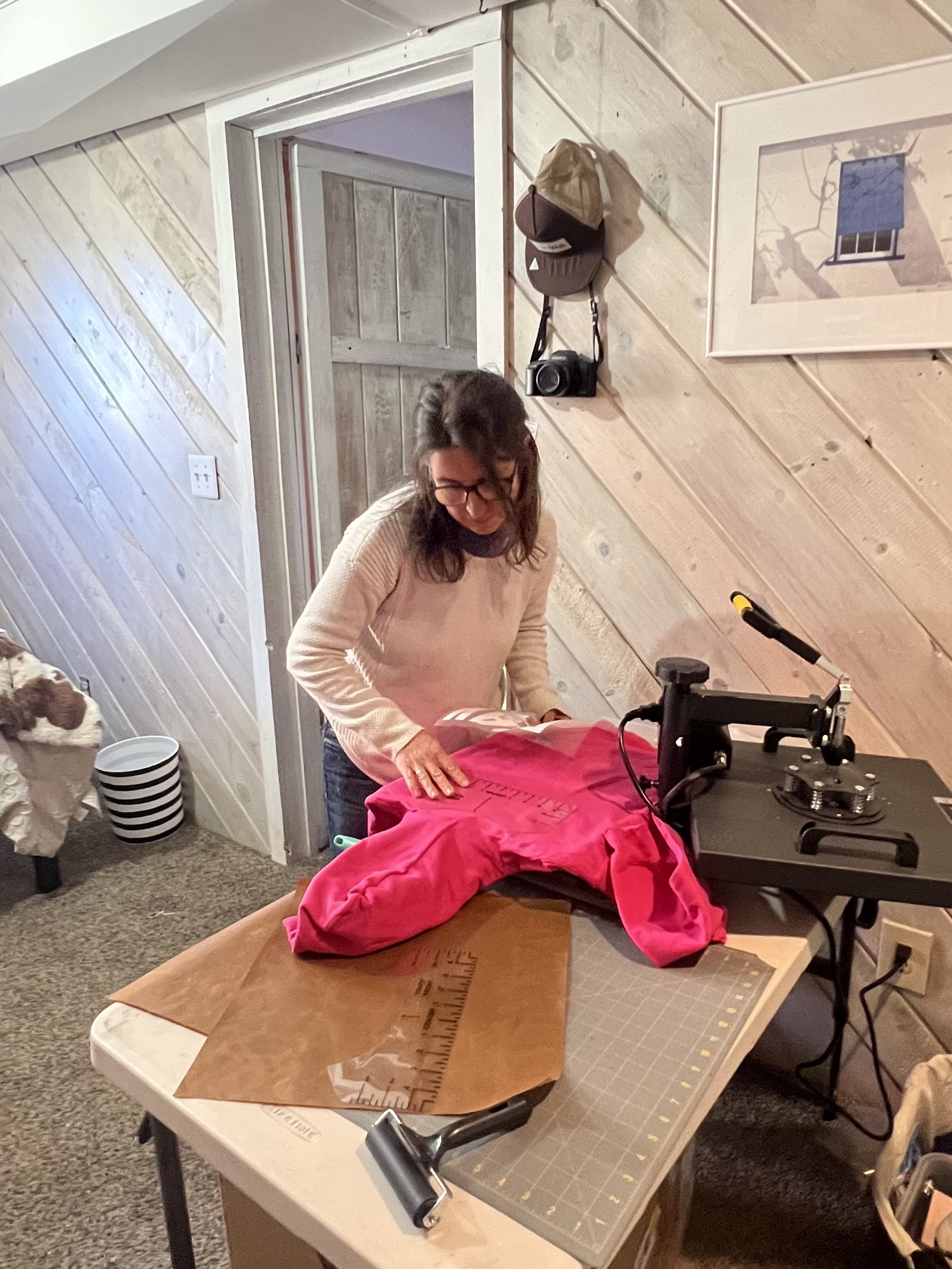 A woman working on a fabric in a workshop, with a table, a cutting mat, and a sewing machine, in a room with wood-paneled walls and a ceiling.