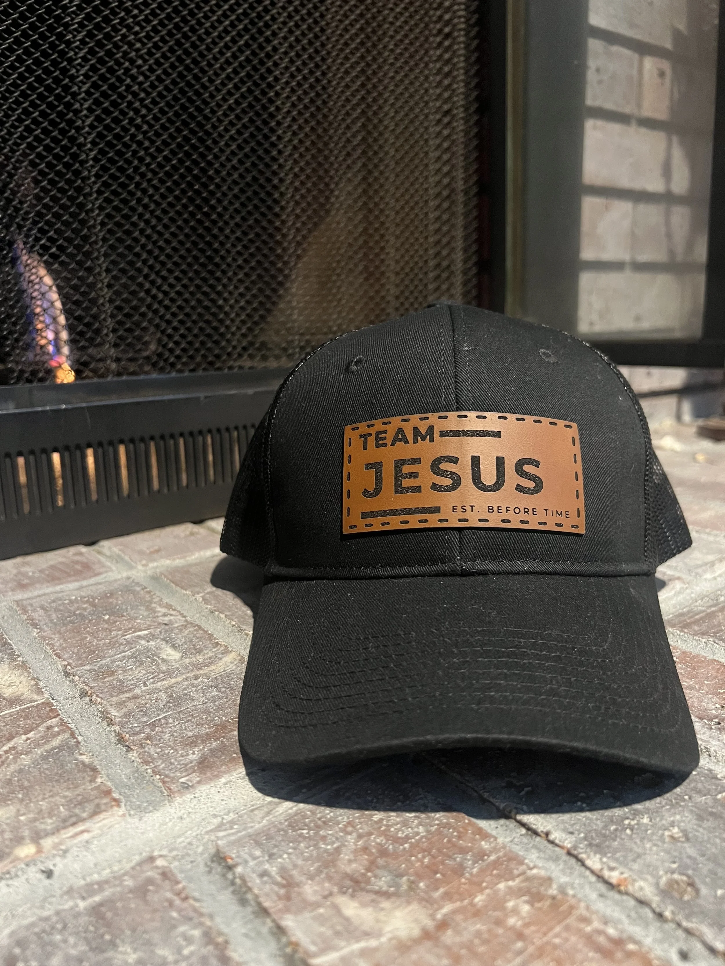 Black baseball cap with a brown patch that reads "Team Jesus - Est. Before Time" placed on brick floor next to a fireplace.