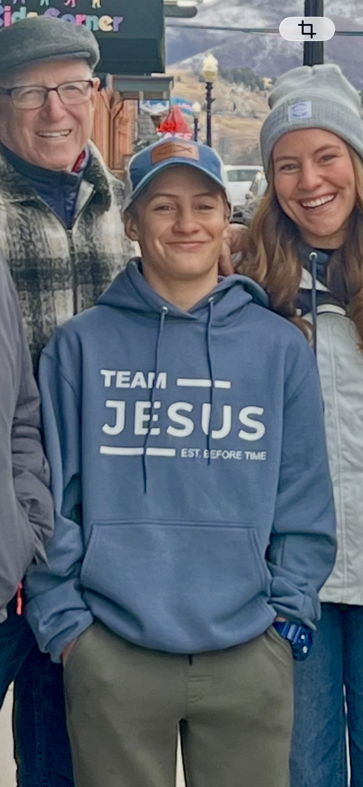 A group of three smiling people, with an older man on the left, a woman on the right, and a young man in the center, wearing a blue hoodie with the text "TEAM JESUS" and a baseball cap, outdoors in a mountainous area.