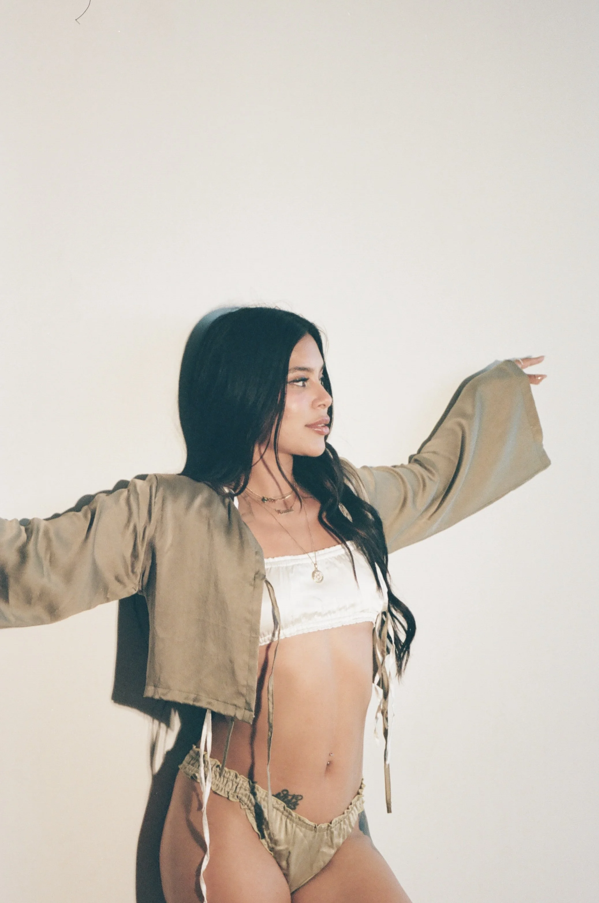 A woman with long dark hair and tattoos wearing a white crop top, beige shorts, and an open beige jacket, standing against a plain white wall with her arms outstretched.