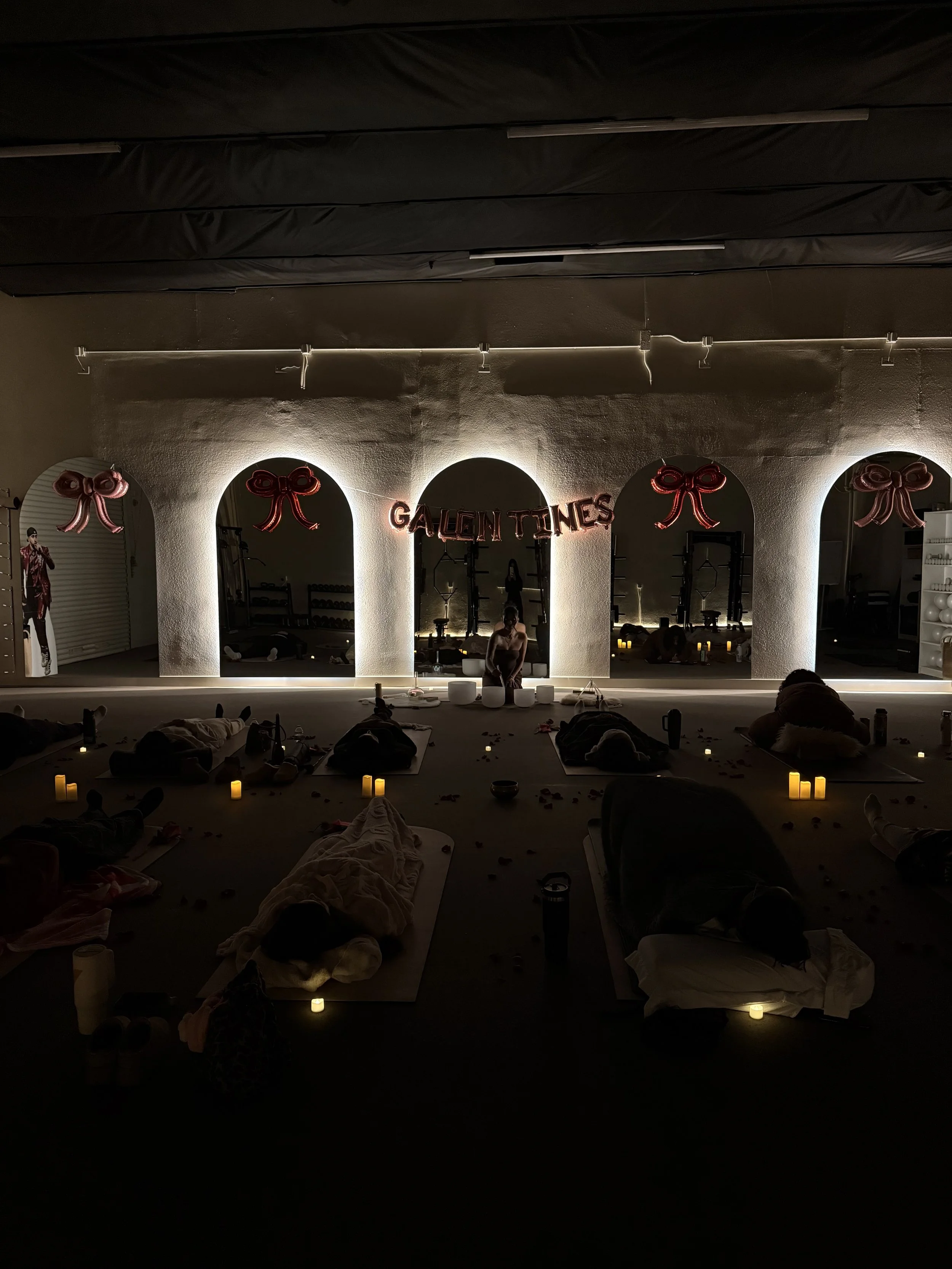 A dimly lit room decorated for a yoga or meditation class with candles and rose-shaped balloons on the walls, and people lying on mats.