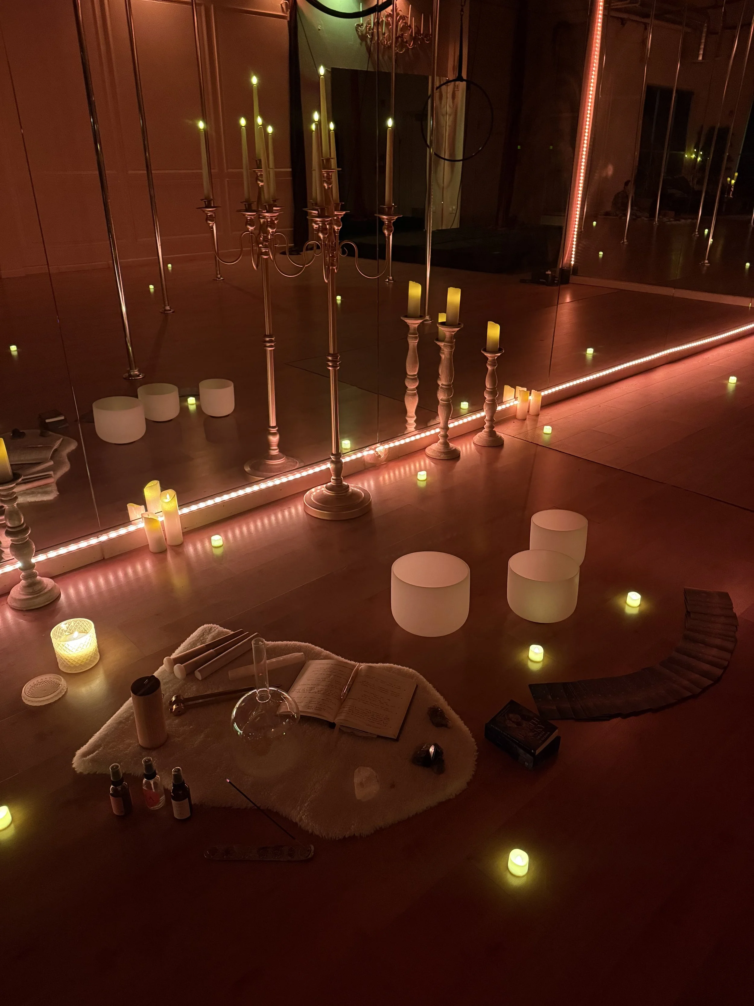 A dimly lit room with a large mirror, candles, and various objects on the floor, including an open book, small bottles, and a glass terrarium, creating a cozy, mystical atmosphere.