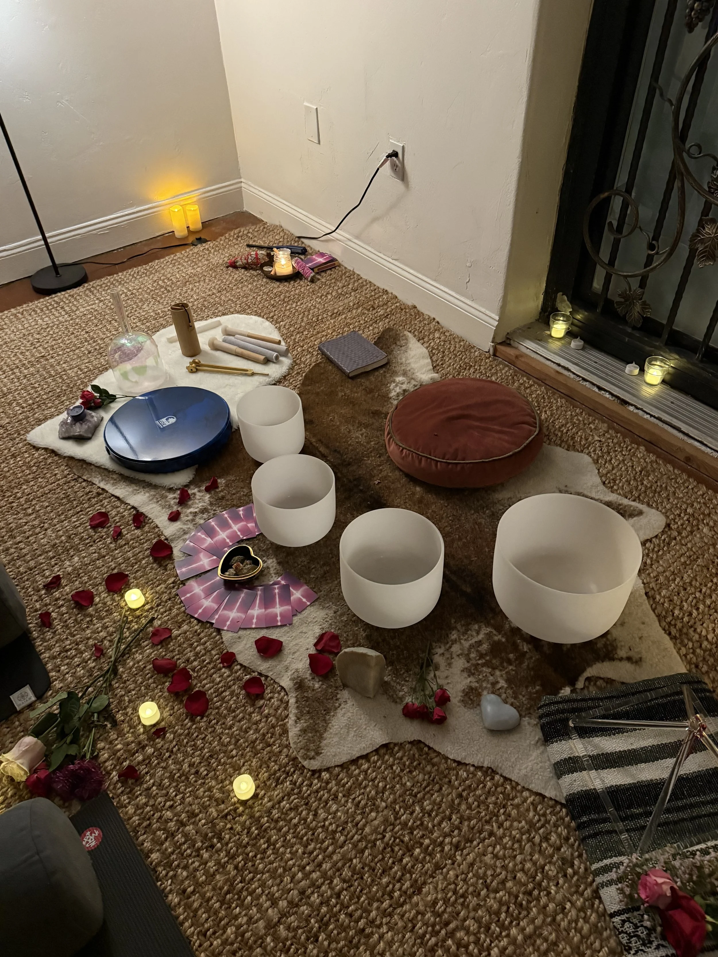 A cozy indoor setting with a tapestry, rose petals, candle lights, singing bowls, a crystal ball, tarot cards, and various objects arranged on a rug, creating an intimate ambiance for a spiritual or meditative session.