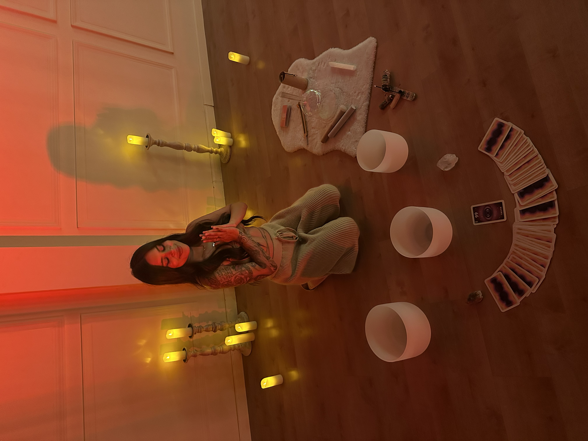 A woman with tattoos on her arms kneeling on the floor in a yoga pose with her hands together in front of her chest, surrounded by candles, tarot cards, crystals, and other spiritual items in a dimly lit room with a warm red-orange glow.