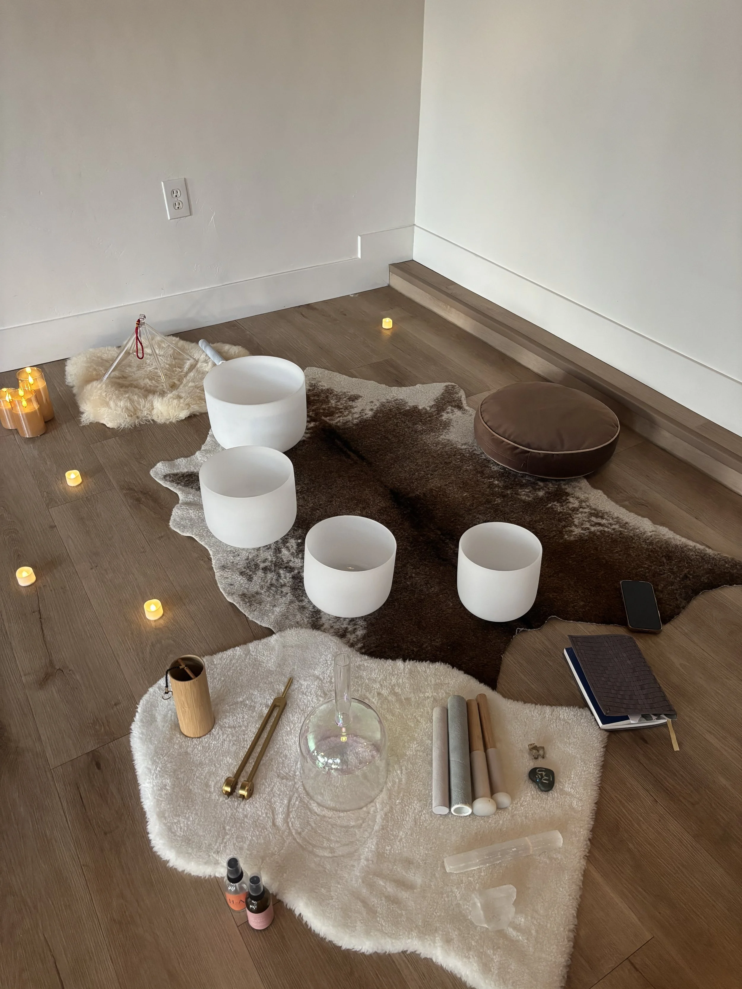 A meditation or yoga setup with white singing bowls on a cowhide rug, candles, crystals, a notebook, and a phone on the wooden floor.