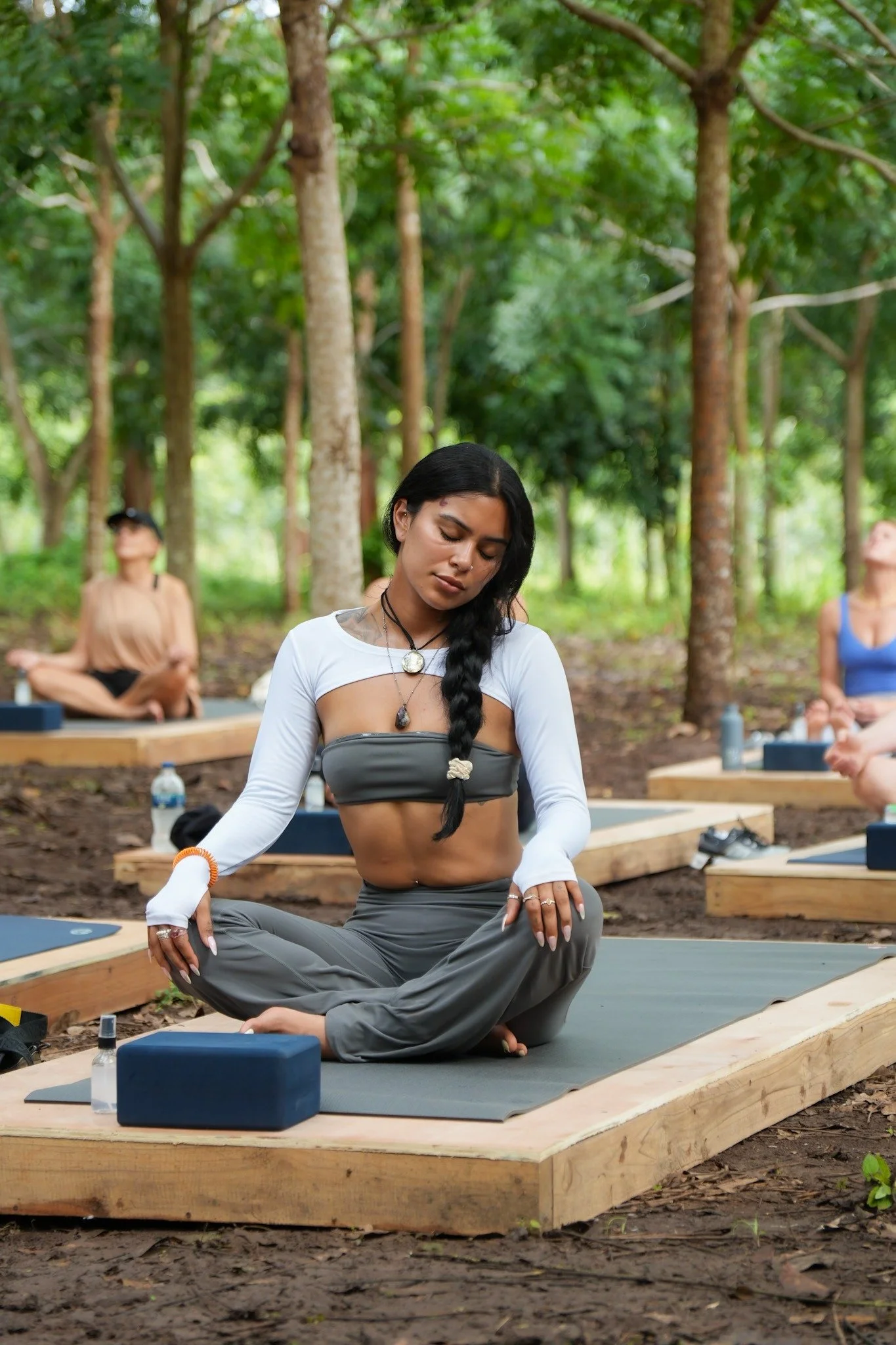 A woman practicing yoga outdoors in a forest, sitting cross-legged on a yoga mat with her eyes closed, surrounded by other people also doing yoga.