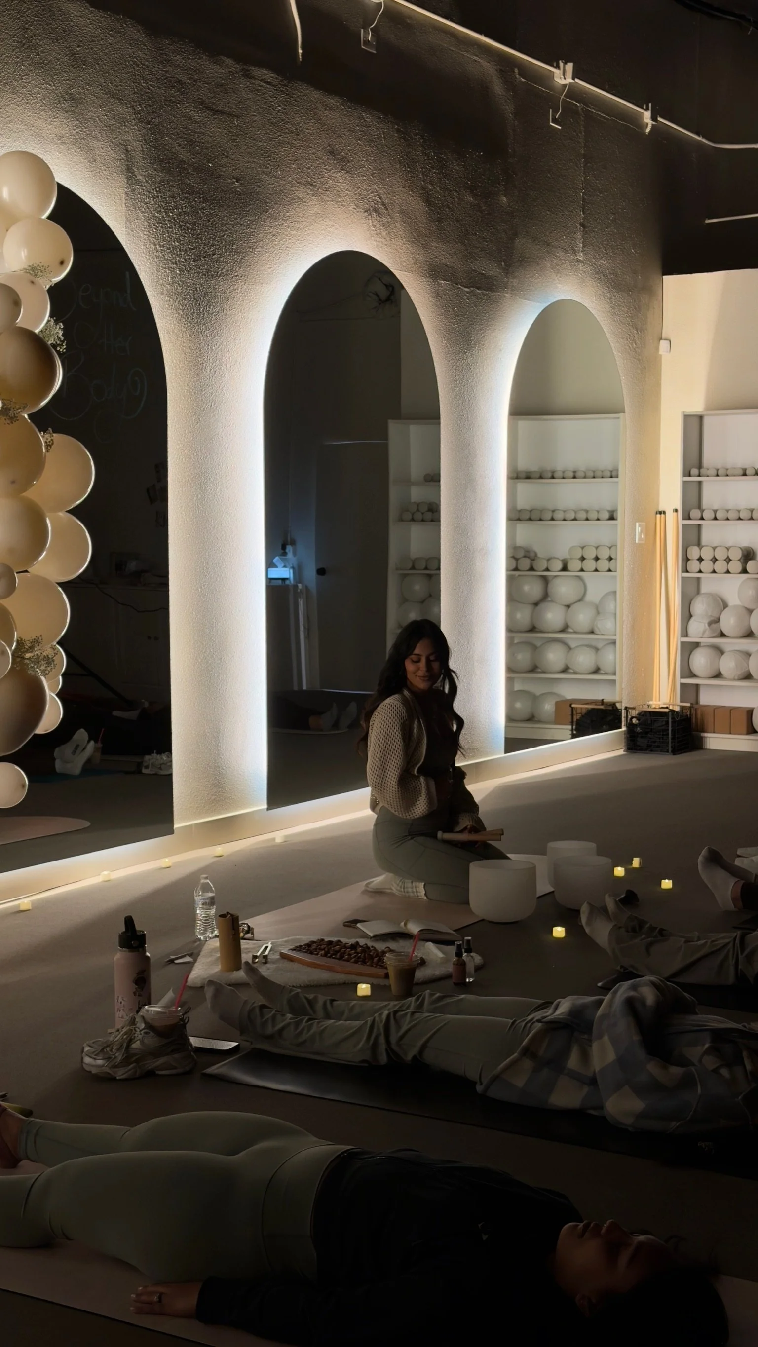 A woman sitting on a yoga mat in a dimly lit room with soft lighting, surrounded by candles. Several people are lying down on yoga mats in a relaxed pose. There are decorative shelves with white spheres in the background.