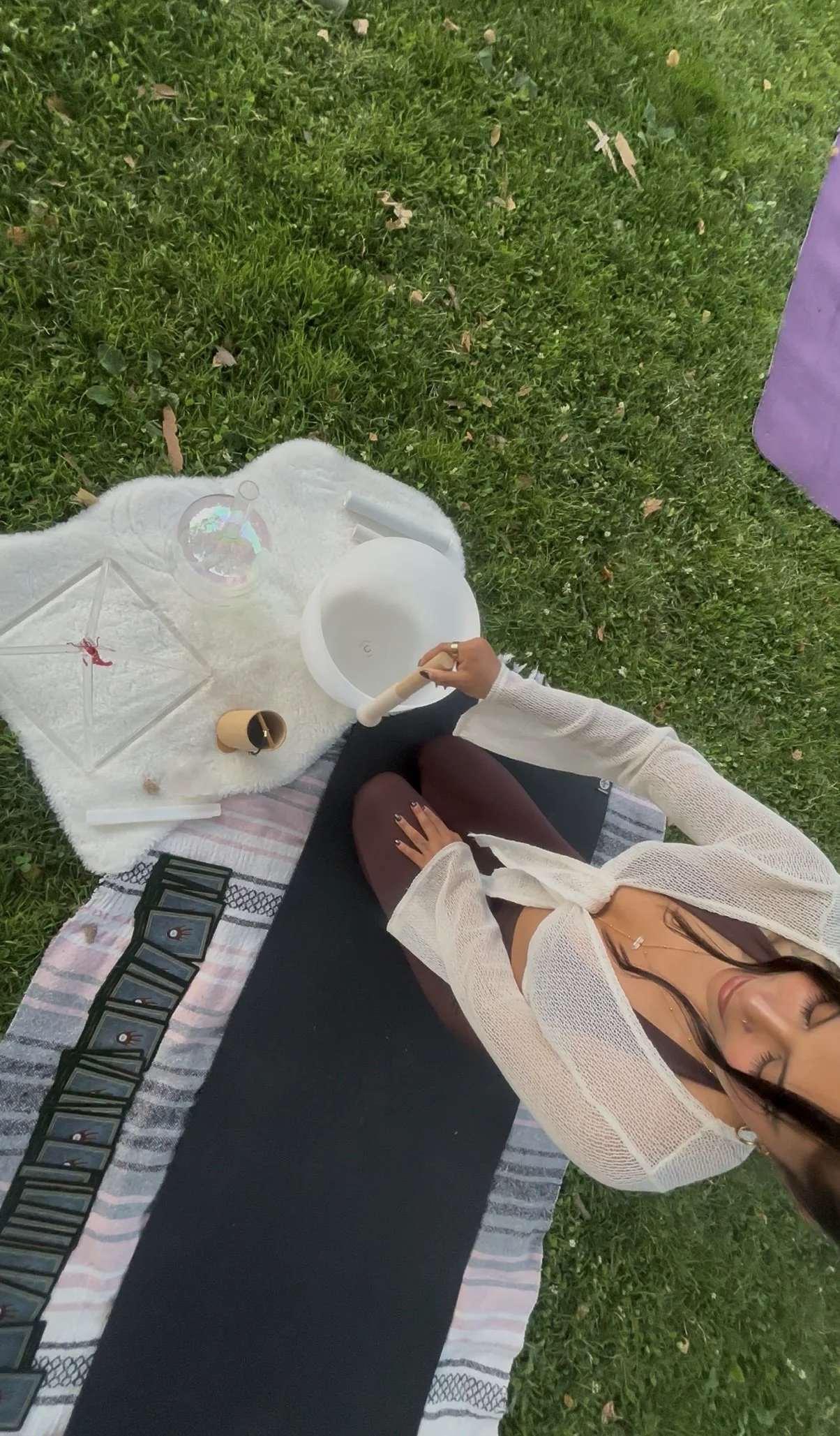 A woman sitting on a striped towel on grass, with a black yoga mat, a white towel, tarot cards, a white bowl, and a clear pyramid-shaped object with a red figurine inside. She is holding a wooden stick or pendulum and appears to be engaged in a spiri