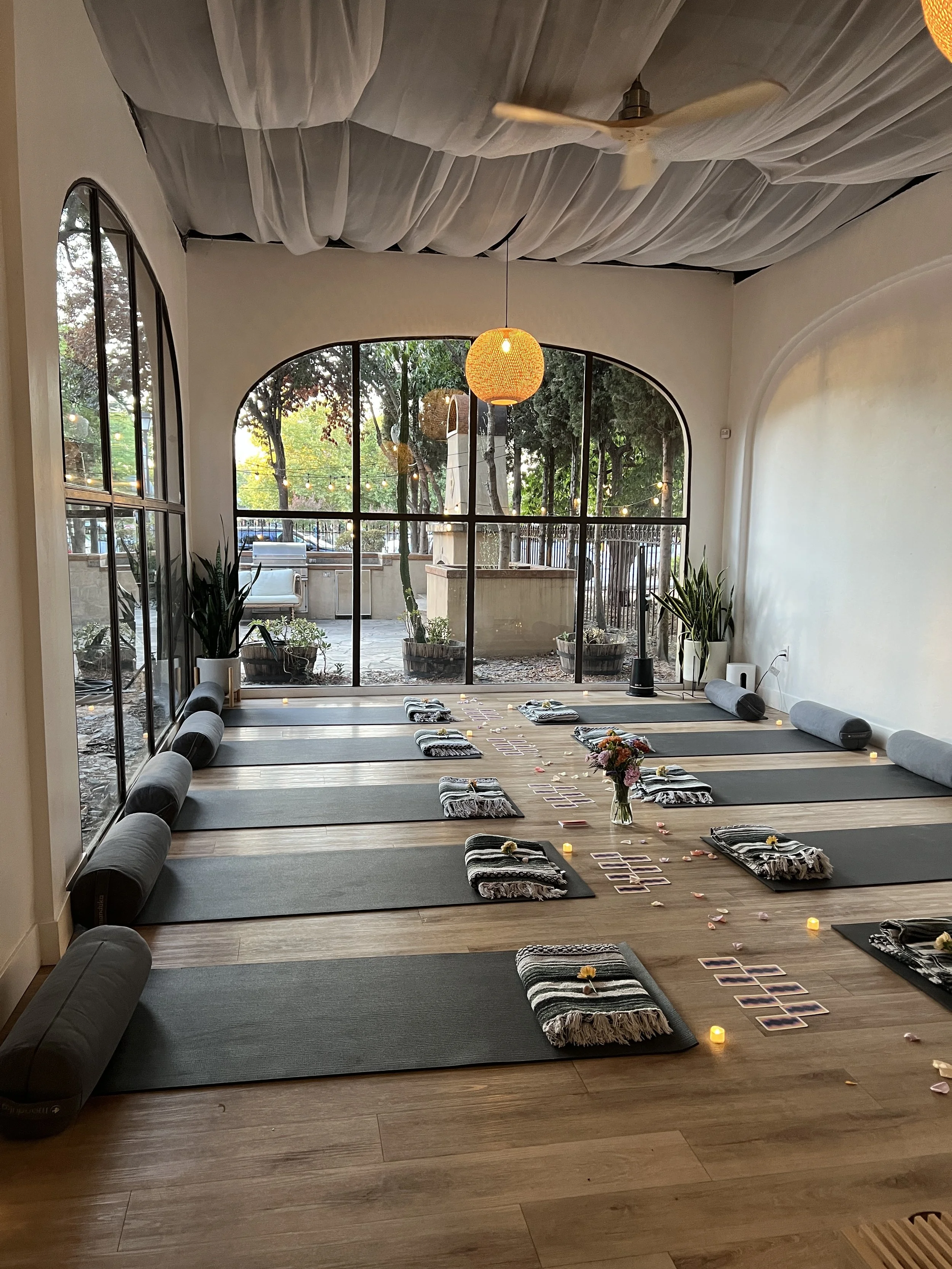 Yoga studio with mats, rolled towels, candles, flowers, and tarot cards arranged for a meditation session, with a large window and outdoor patio visible.