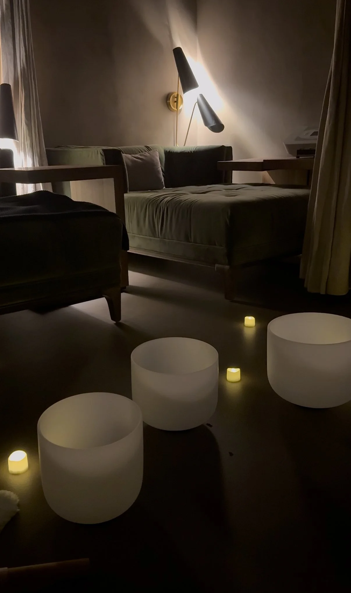 A cozy living room scene with a green sofa, a wooden side table, and a wall-mounted lamp with three adjustable black arms. There are white glow-in-the-dark orbs on the floor, casting a gentle light in the dimly lit room.
