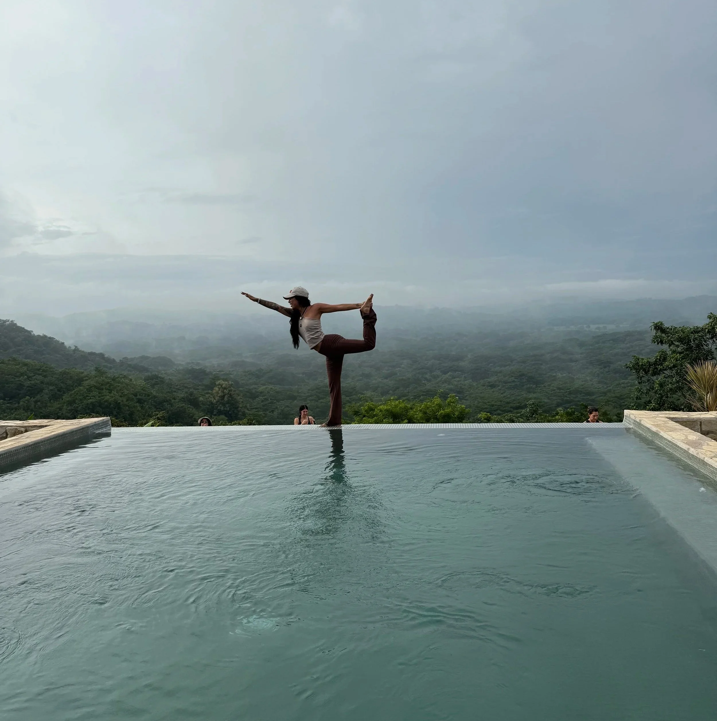 A woman performing a yoga pose on the edge of an infinity pool with lush green hills and cloudy sky in the background.
