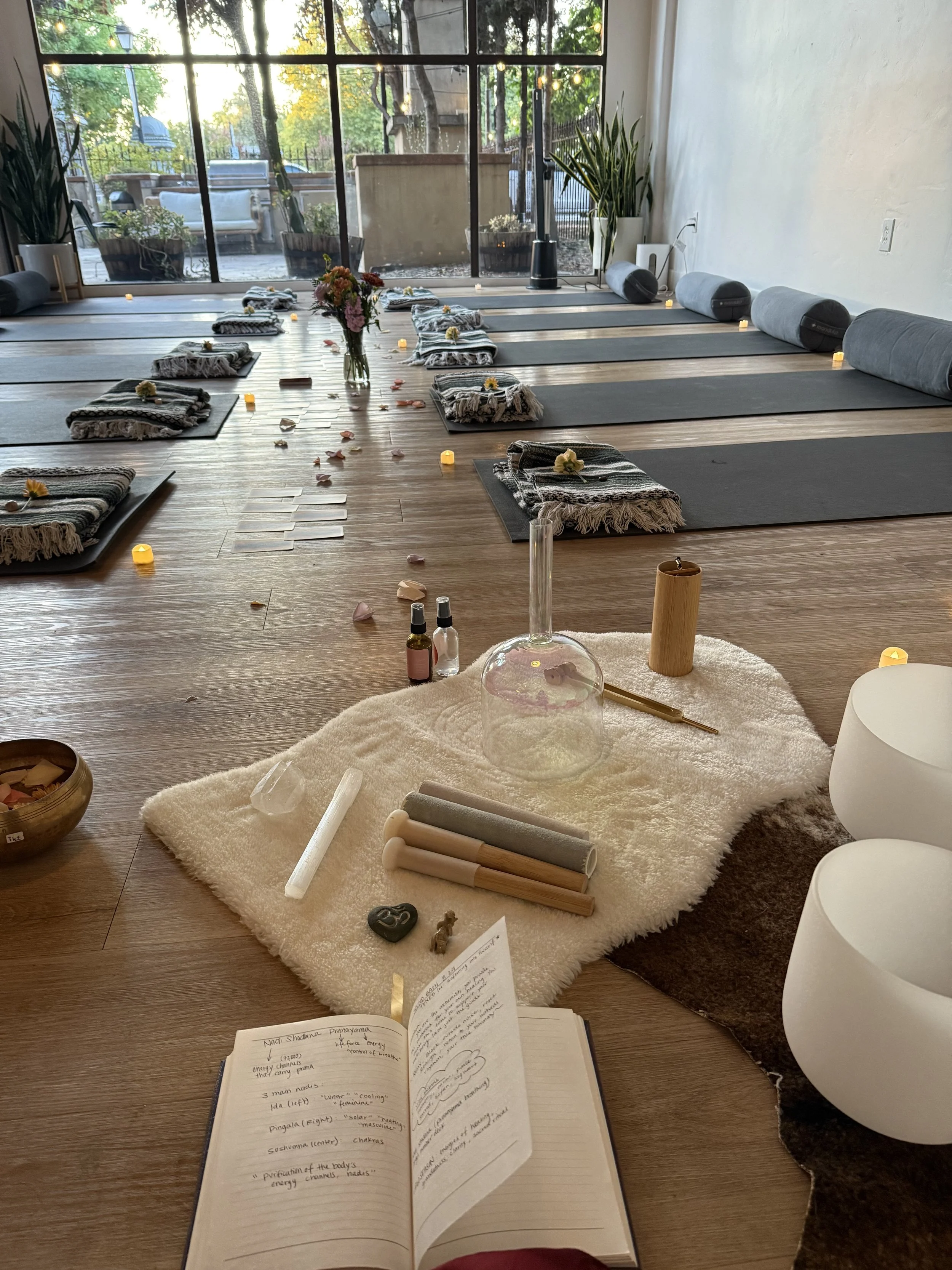 A yoga or meditation studio with mats and bolsters arranged for a class. The room has large windows with a view of trees outside. On the floor in the foreground, there are candles, small bottles, a notebook with handwritten notes, and various ritual 
