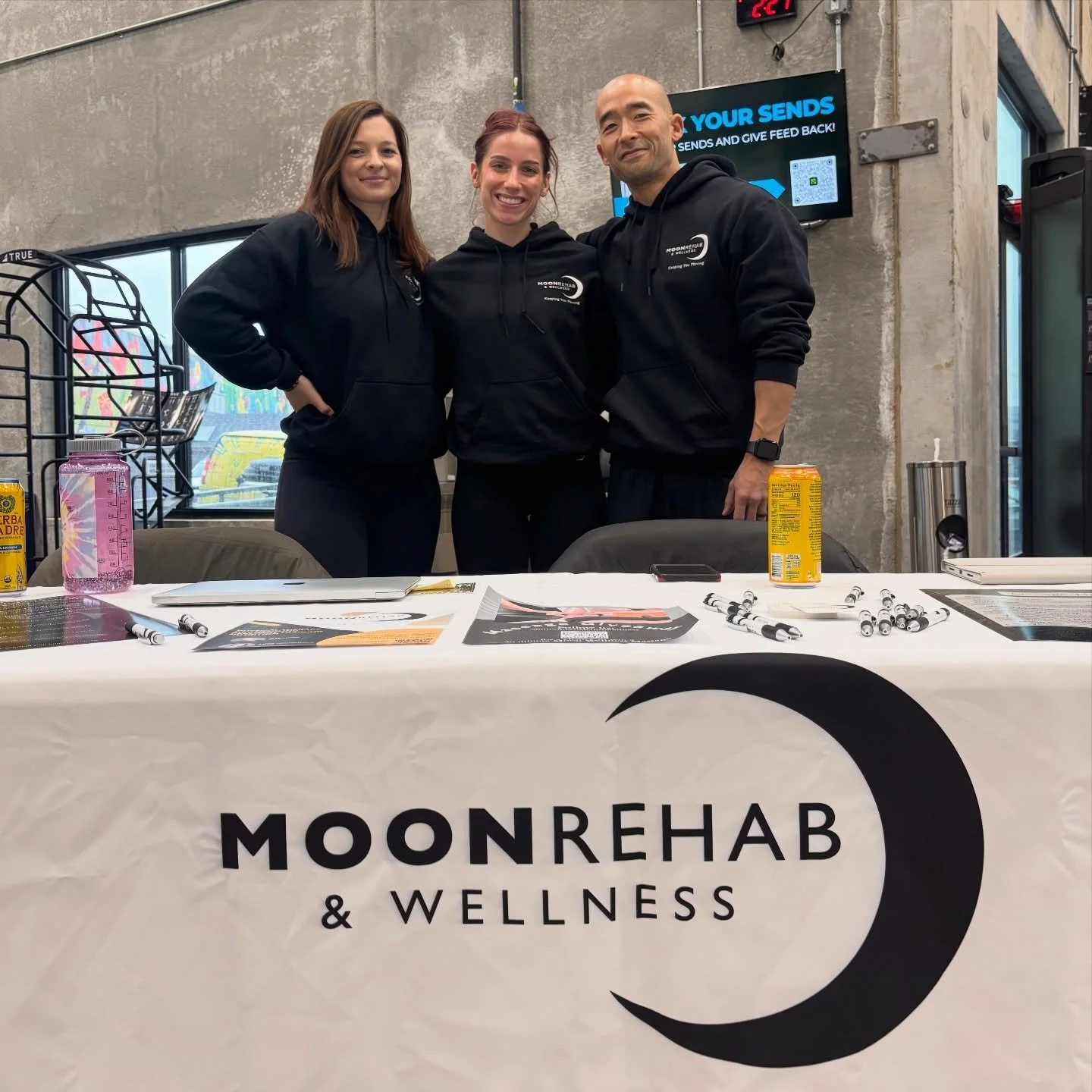 Three people standing behind a table at a wellness event, all wearing black hoodie with 'MOONREHAB & WELLNESS' logos. The table has pens, flyers, a yellow drink, and a water bottle. Behind them, a concrete wall and a digital screen display information. The table has a cloth with the company logo, featuring a crescent moon symbol.
