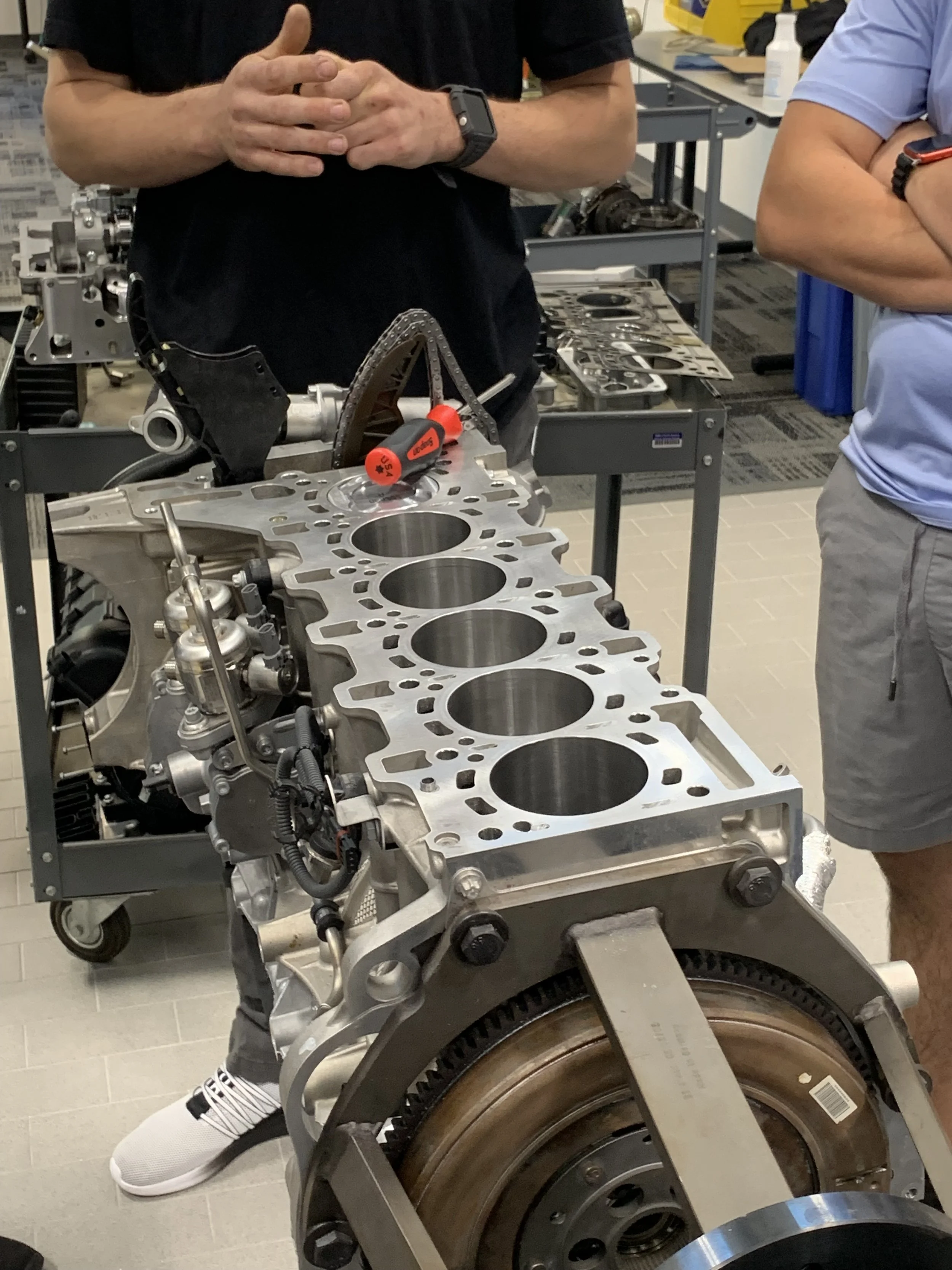 People examining a disassembled engine block and cylinder heads in a workshop, with tools and equipment in the background.