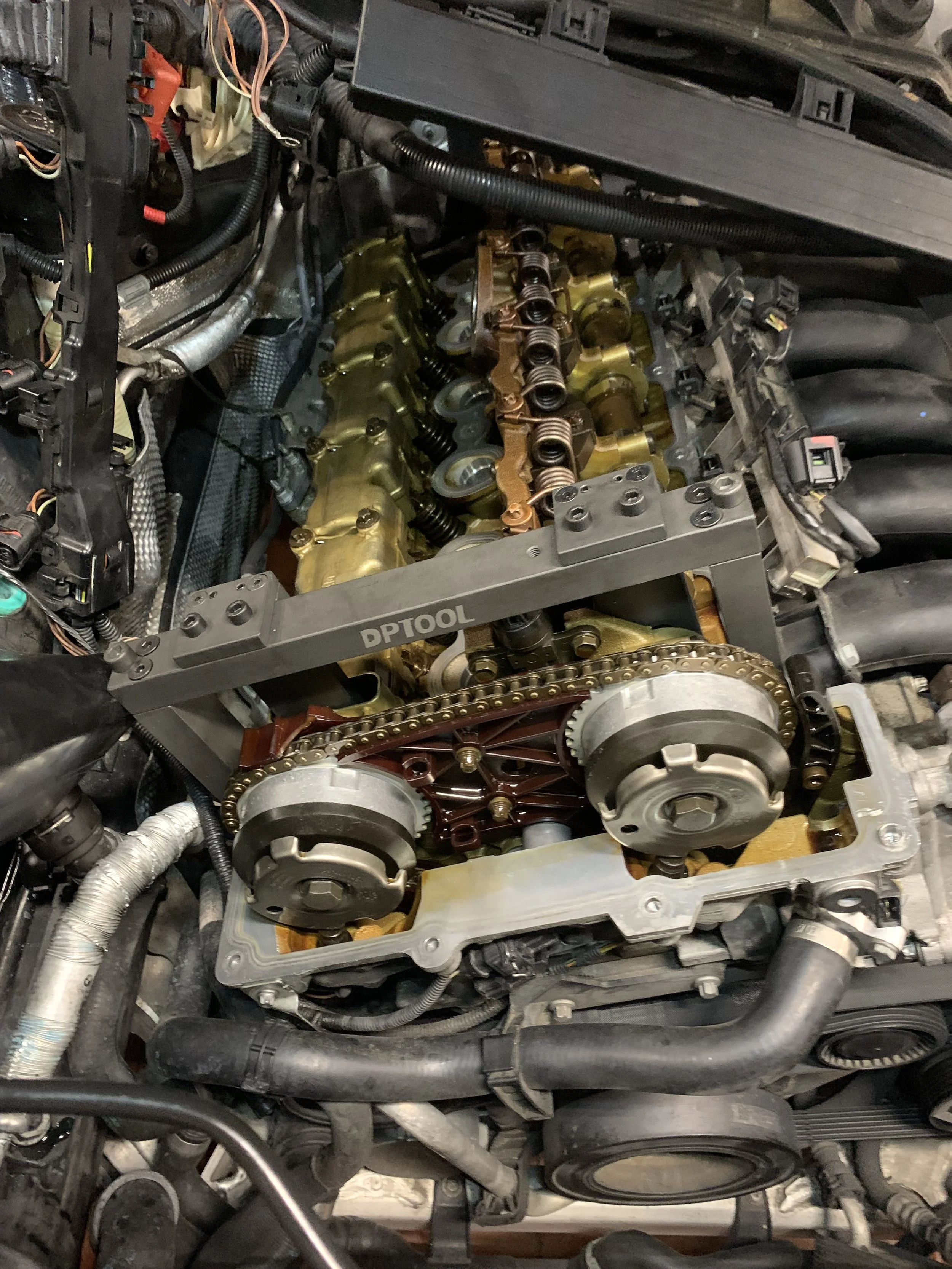 Close-up of a car engine during repair, showing the valve cover removed, exposing the camshaft and timing chain, with various engine components and wiring visible.