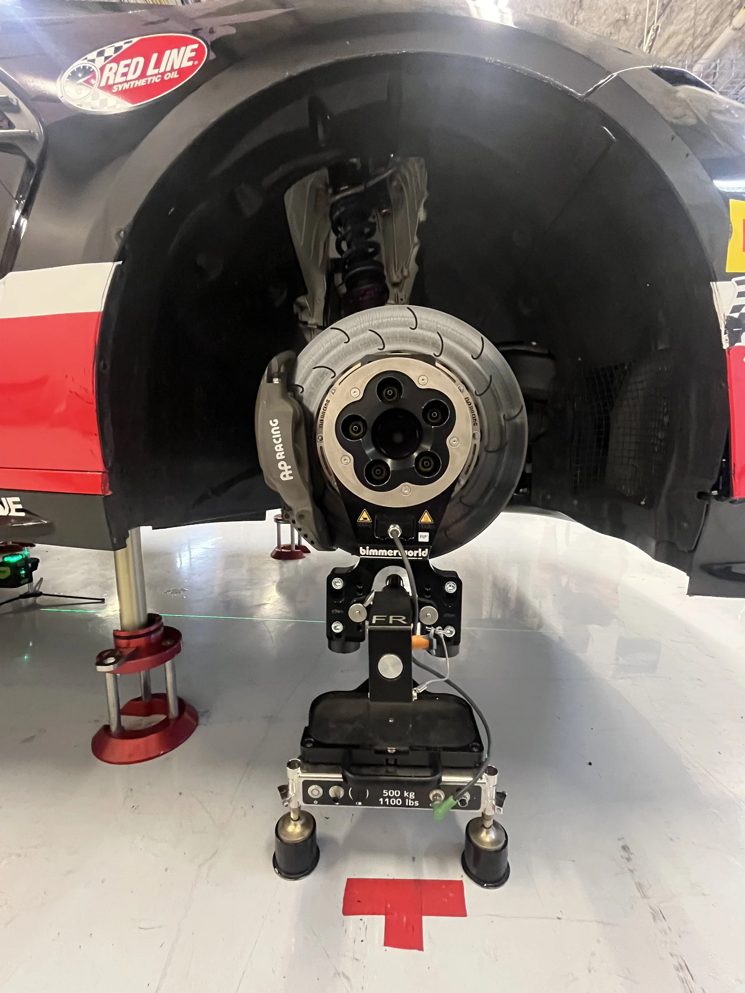 Race car wheel and brake system being tested on a dynamometer, with support equipment and red markings on the floor.