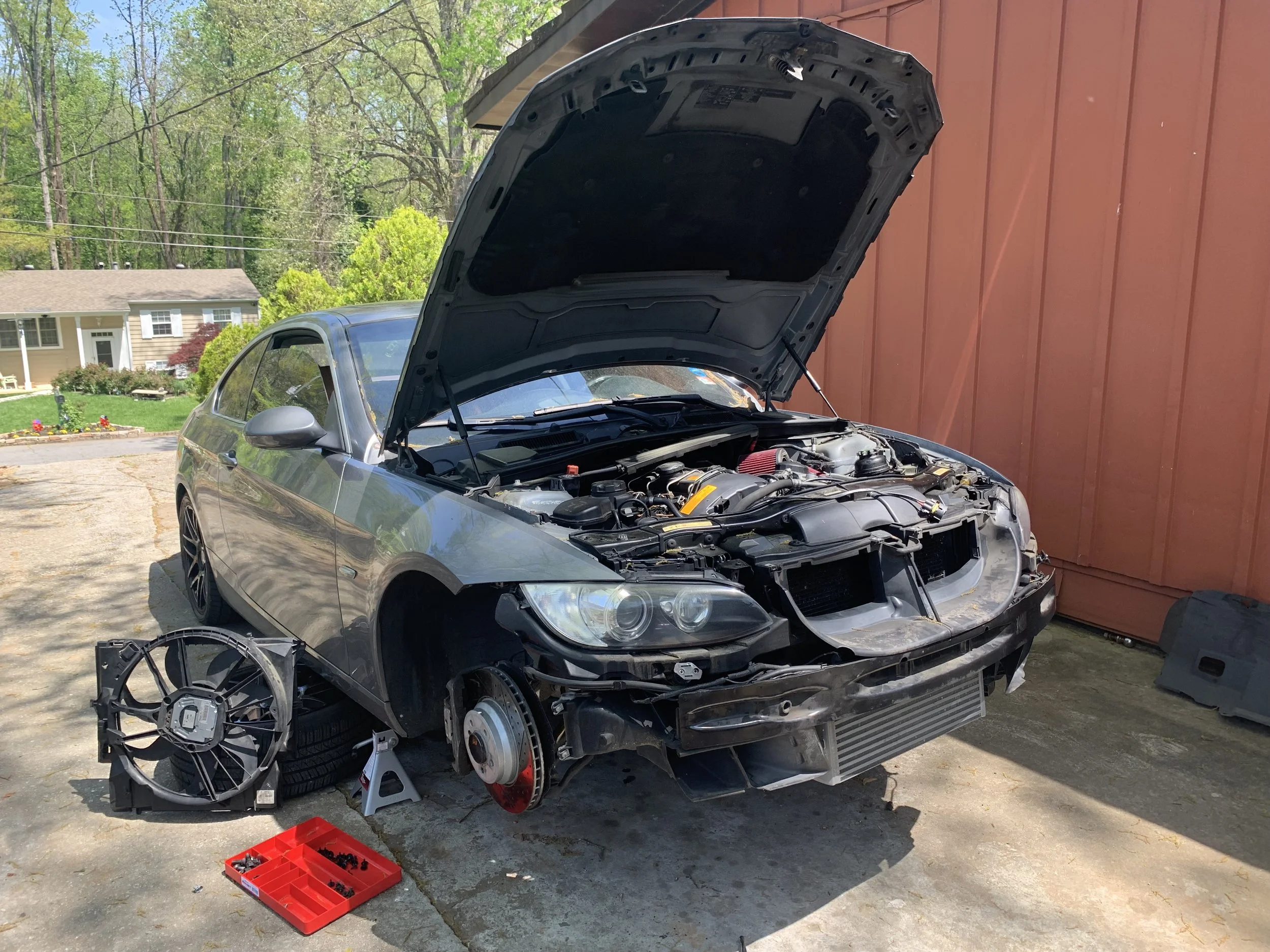 A silver sports car with the front bumper removed is parked outside near a red building, with its hood open and a detached wheel lying on the ground nearby. A red parts tray with various small parts is on the ground in front of the car.