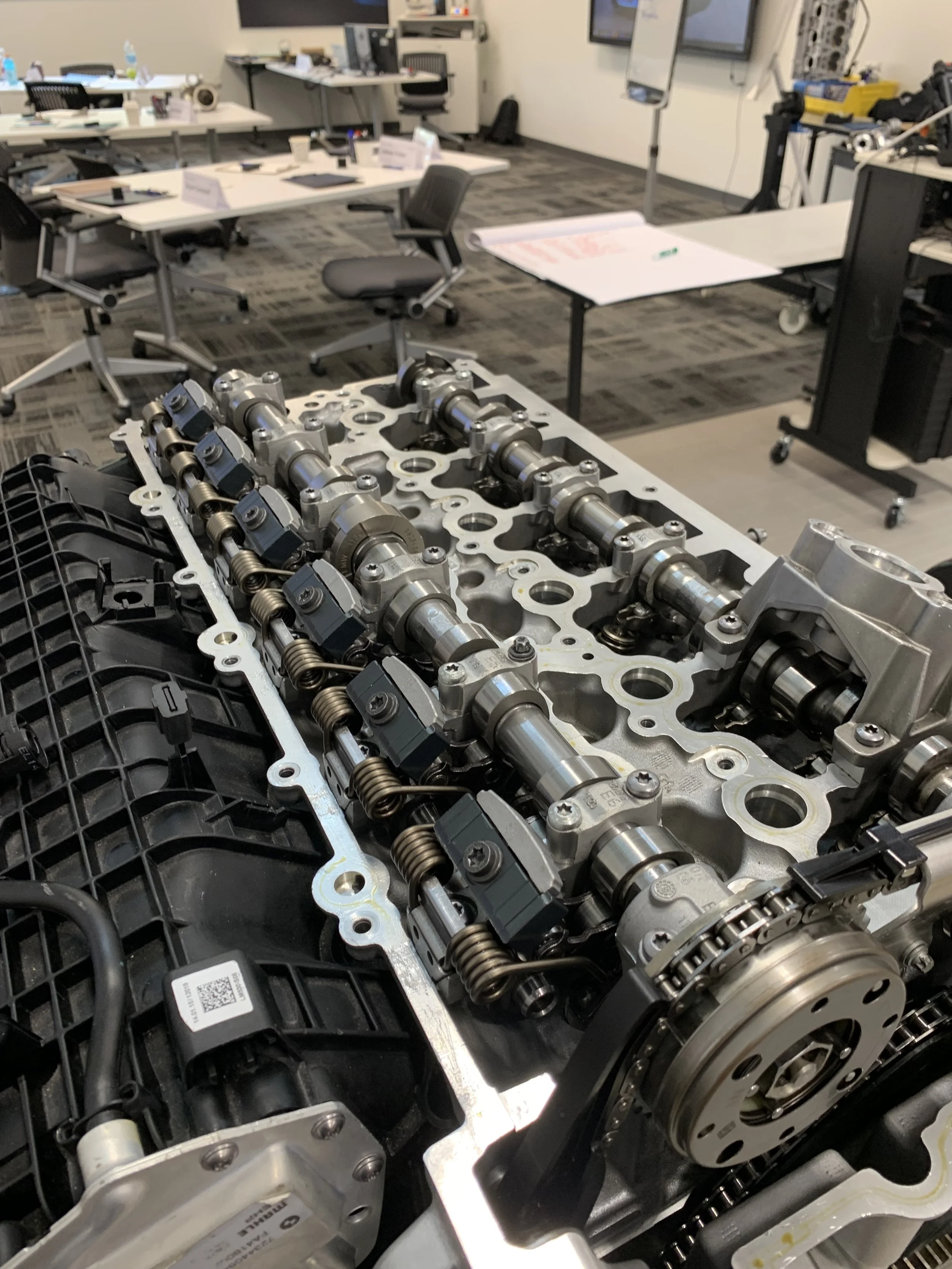 Close-up of an automotive engine component, specifically the camshaft with various mechanical parts surrounding it, in a professional workshop or office setting.