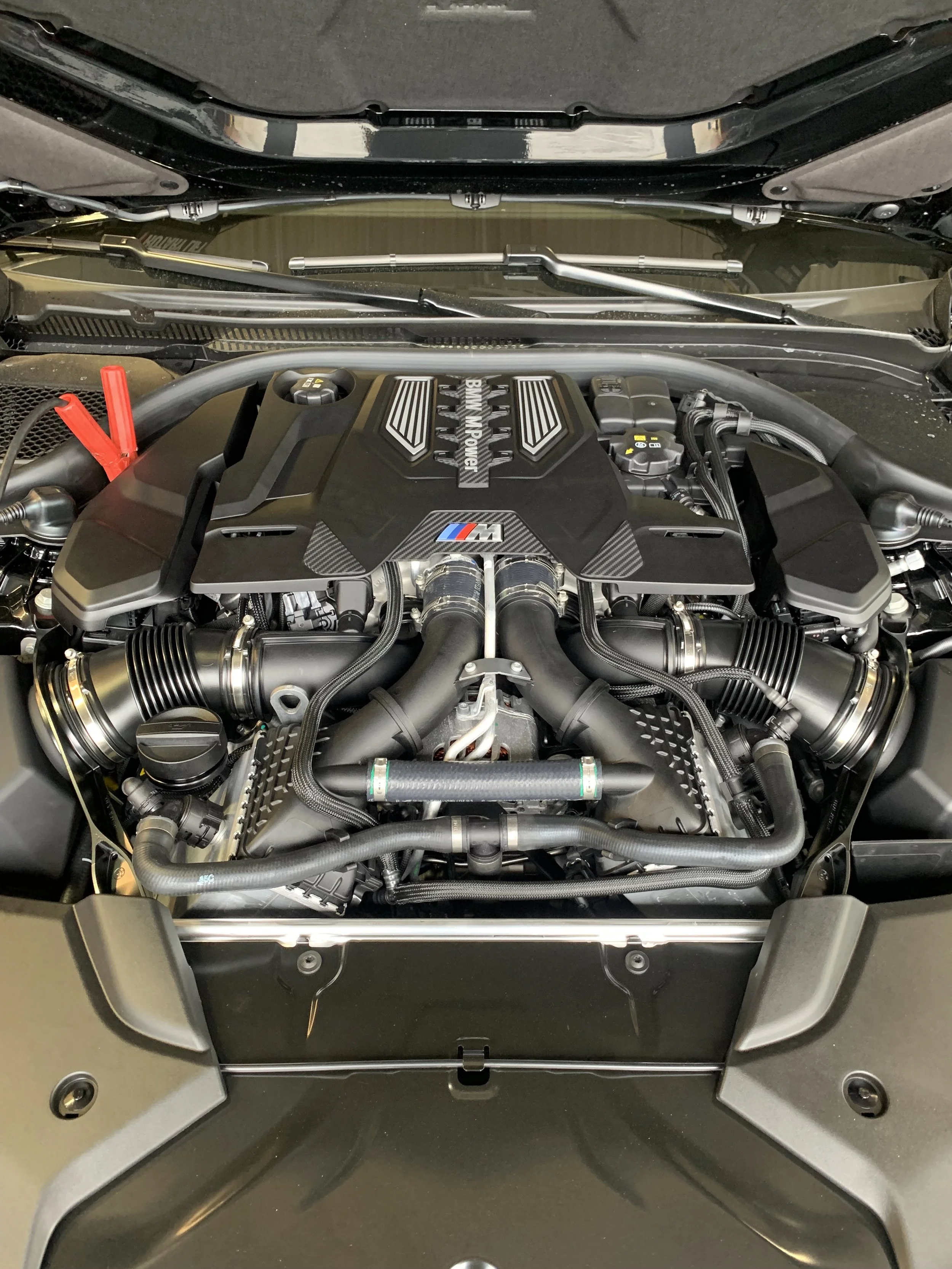 Close-up of a high-performance car engine with BMW M branding, featuring various pipes, hoses, and mechanical components.