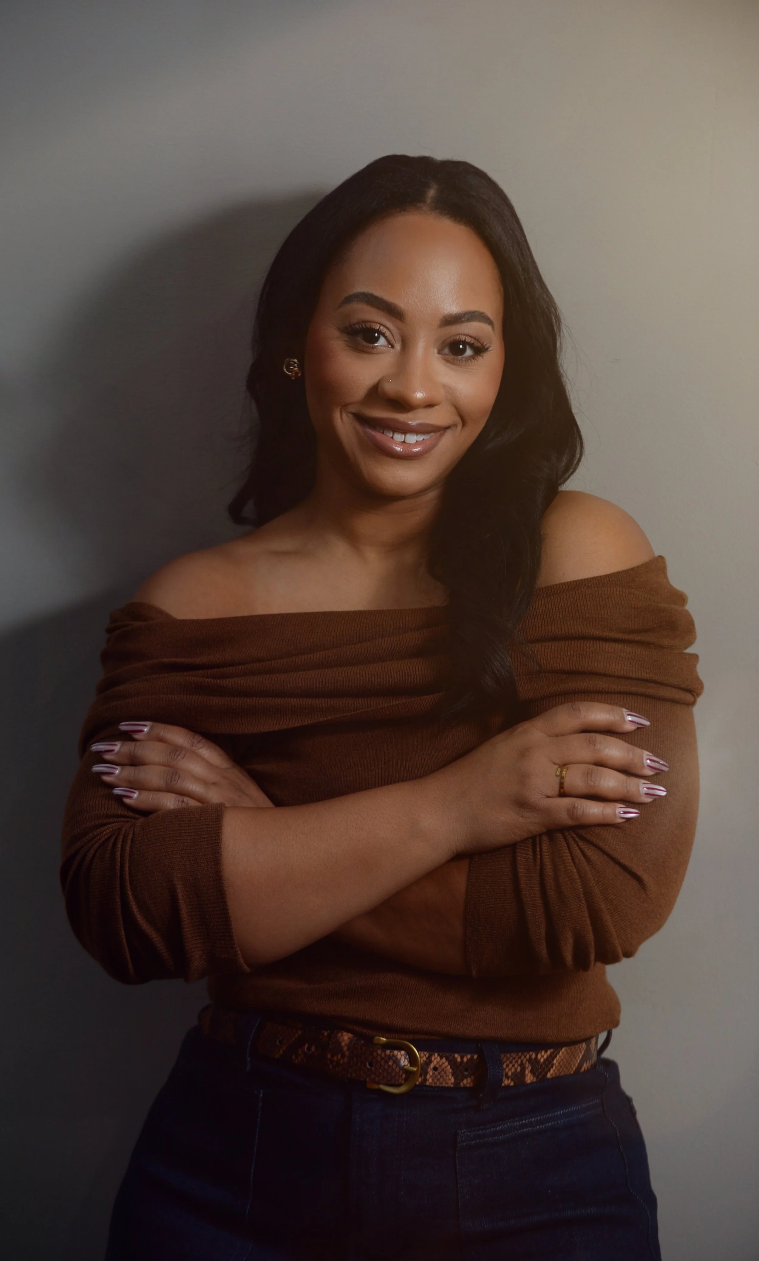 A woman with dark hair, smiling, wearing a brown off-the-shoulder top and dark jeans, crossing her arms, standing against a plain wall.