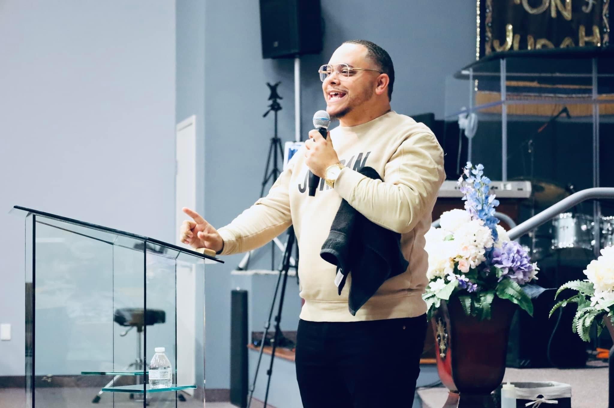 A man wearing glasses and a beige sweatshirt speaking into a microphone during a presentation or speech. He is smiling and gesturing with his right hand. There is a table with a water bottle in front of him, and a flower arrangement next to the stage.