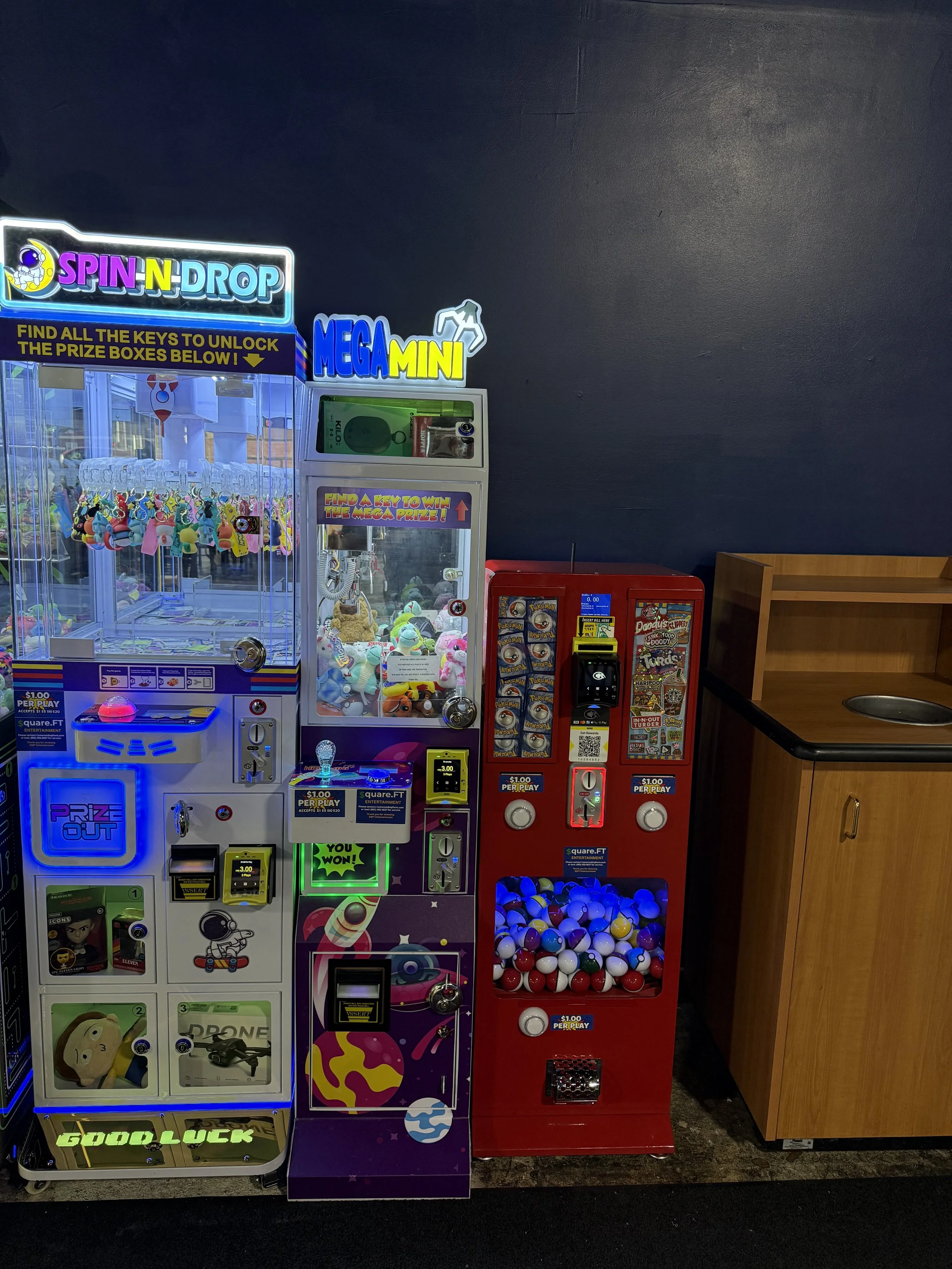 Two arcade claw machines are side by side, filled with plush toys and prizes. The machine on the left is themed 'Spin N Drop' with bright lights, colorful plush toys inside, and a sign advertising prize boxes. The right machine is a red claw game filled with plastic balls, with bright signage and a dollar per play fee. Next to the arcade machines is a wooden cabinet with a black countertop and a metal circular sink.