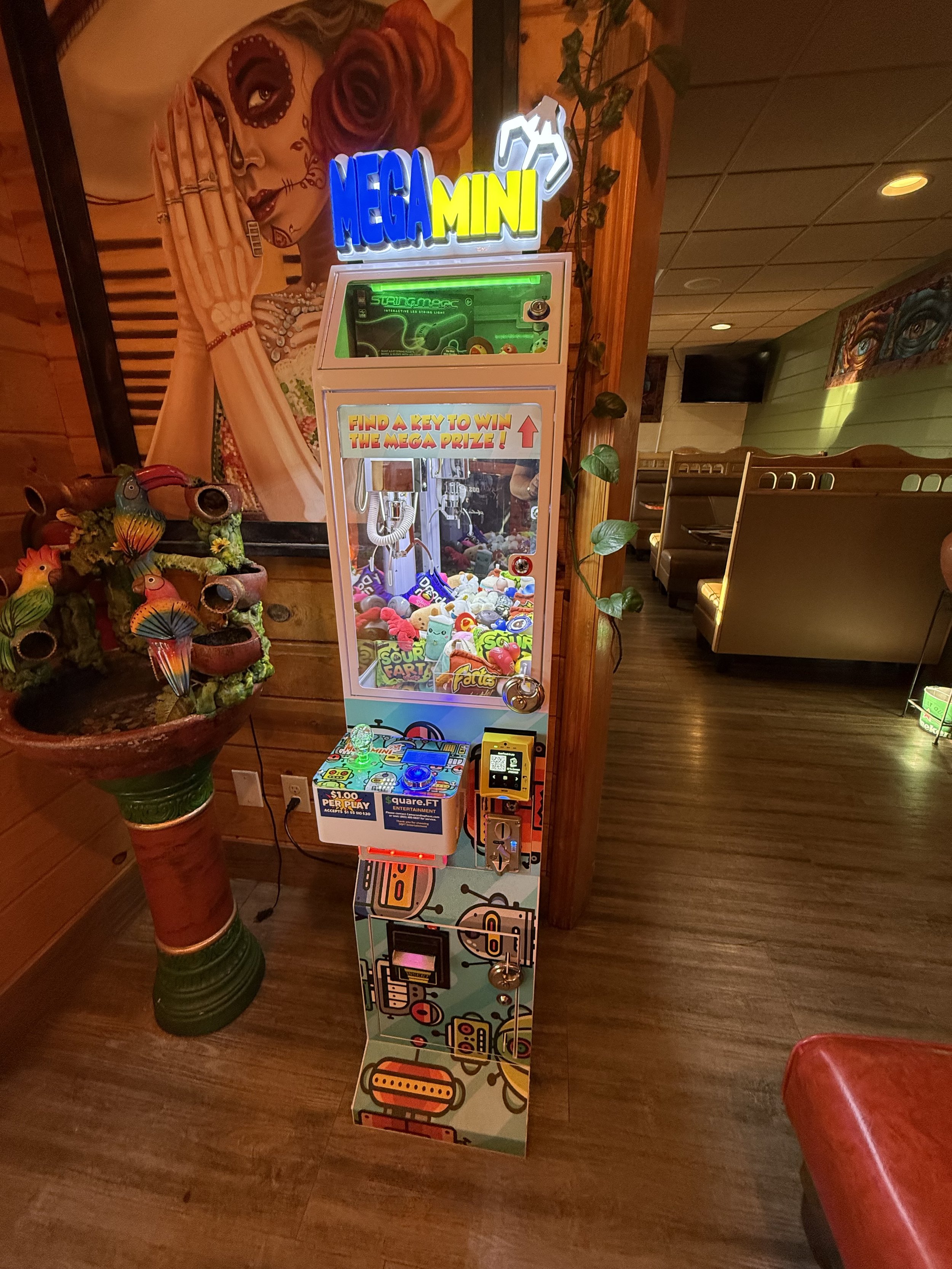 A colorful claw machine game called 'Mega Mini' filled with plush toys and prizes, with a sign that says 'Find a key to win the mega prize.' The machine has a digital screen, coin slot, and a joystick for control, situated in a lively restaurant or arcade setting with wall art and booths in the background.