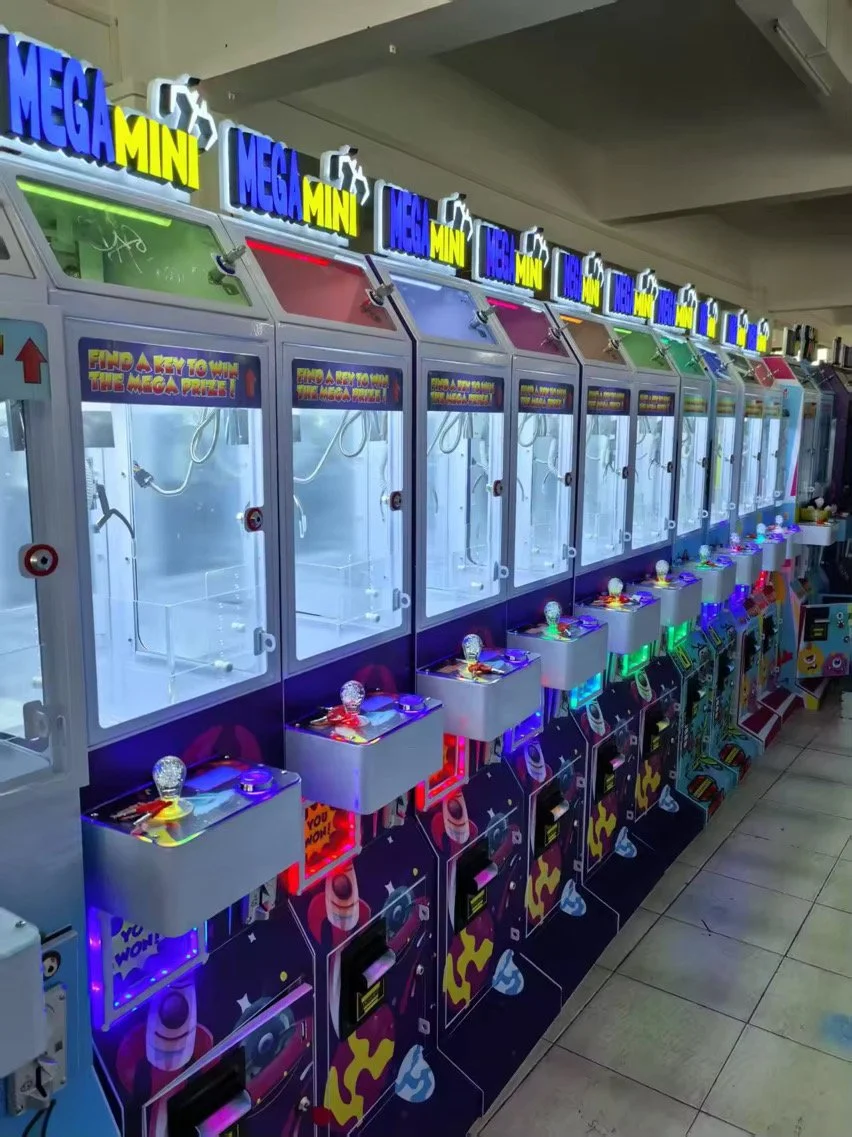 A row of colorful arcade claw machines labeled 'Mega Mini' with illuminated signs on top, each with a visible glass enclosure and a control panel with a doll light at the top, located inside an arcade or entertainment center.
