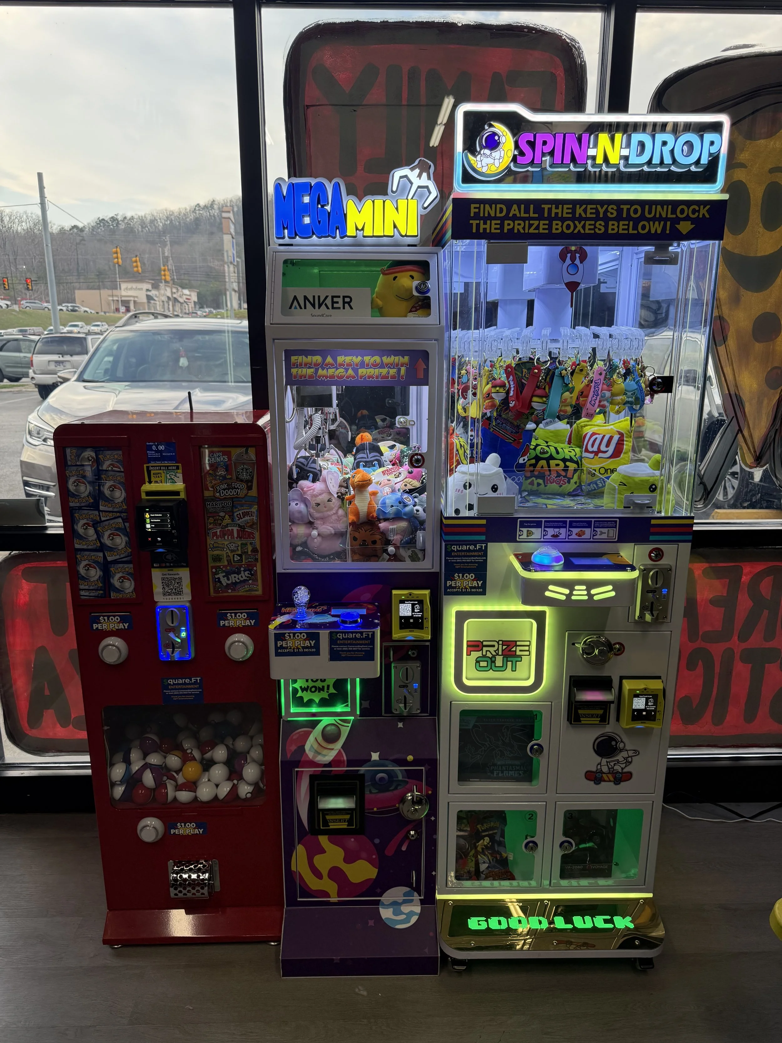 A collection of arcade claw prize machines inside a store, with a window showing parked cars outside.