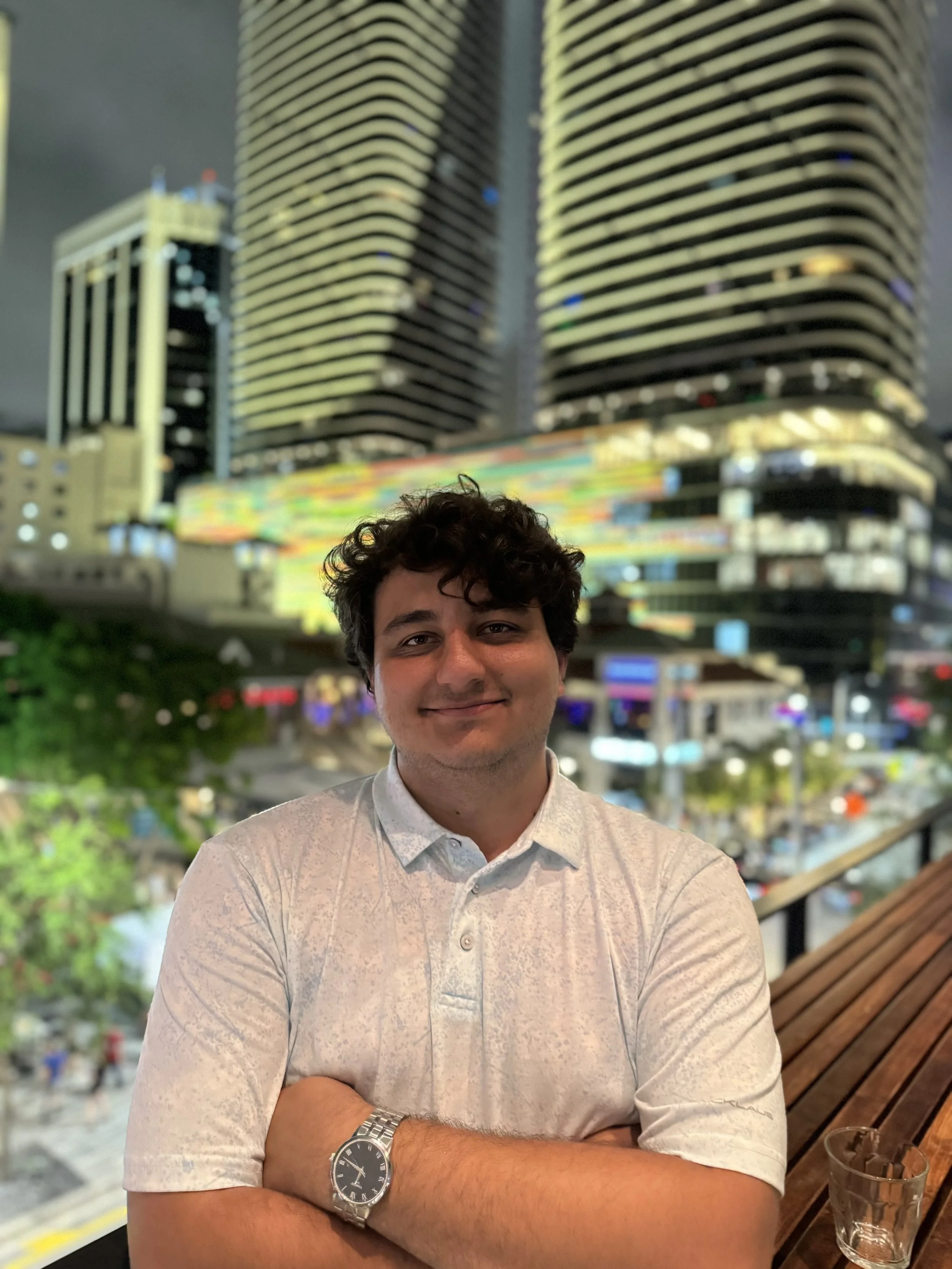 A man with dark curly hair and a light-colored shirt stands with arms crossed, smiling, against a background of tall buildings with colorful lights at night.