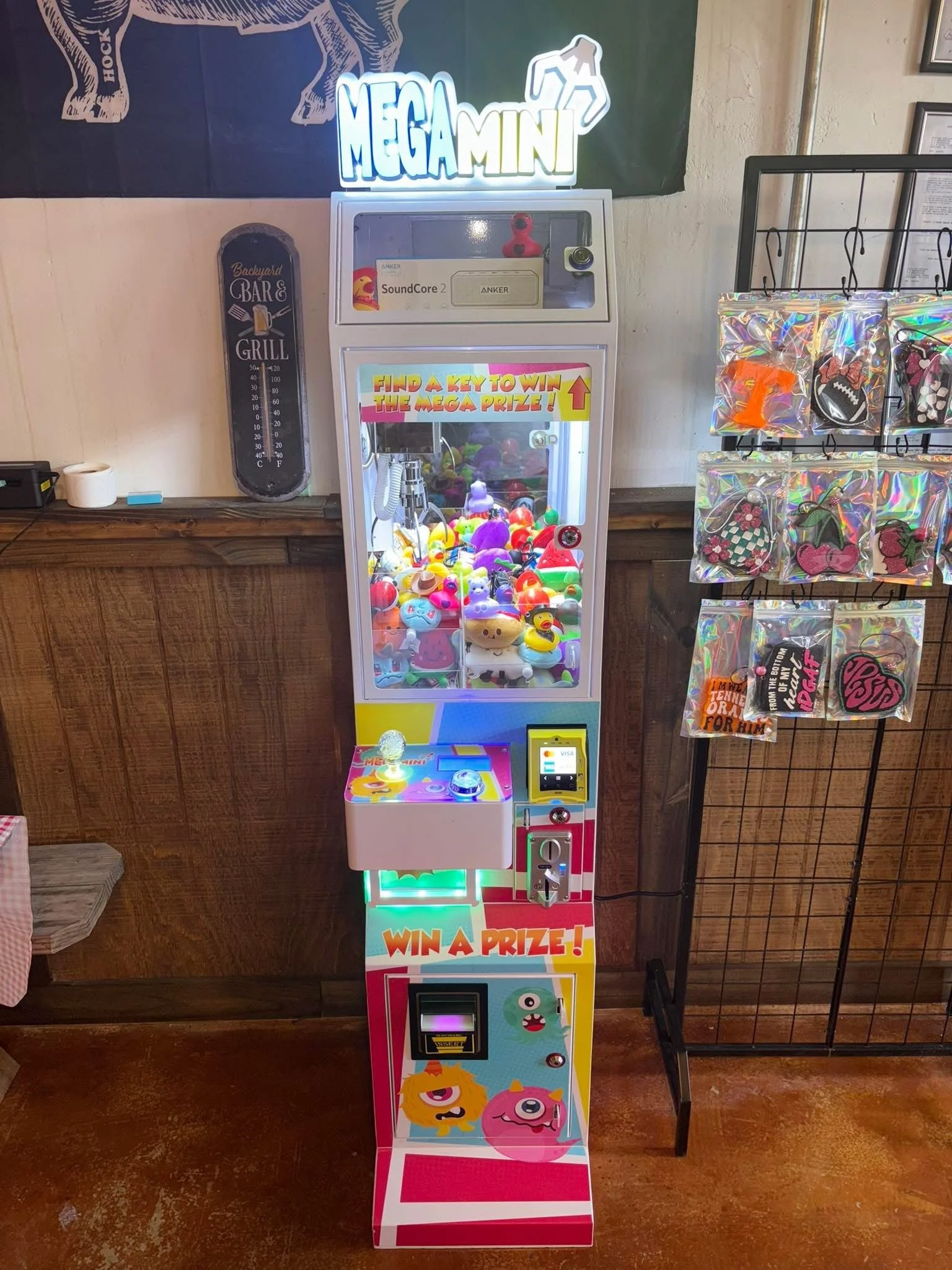 Colorful claw machine arcade game labeled 'MegaMini' with a sign above that says 'Find a key to win the mega prize.' It has various plush toys inside and a sticker at the bottom with cartoon monsters and a slot to win a prize.