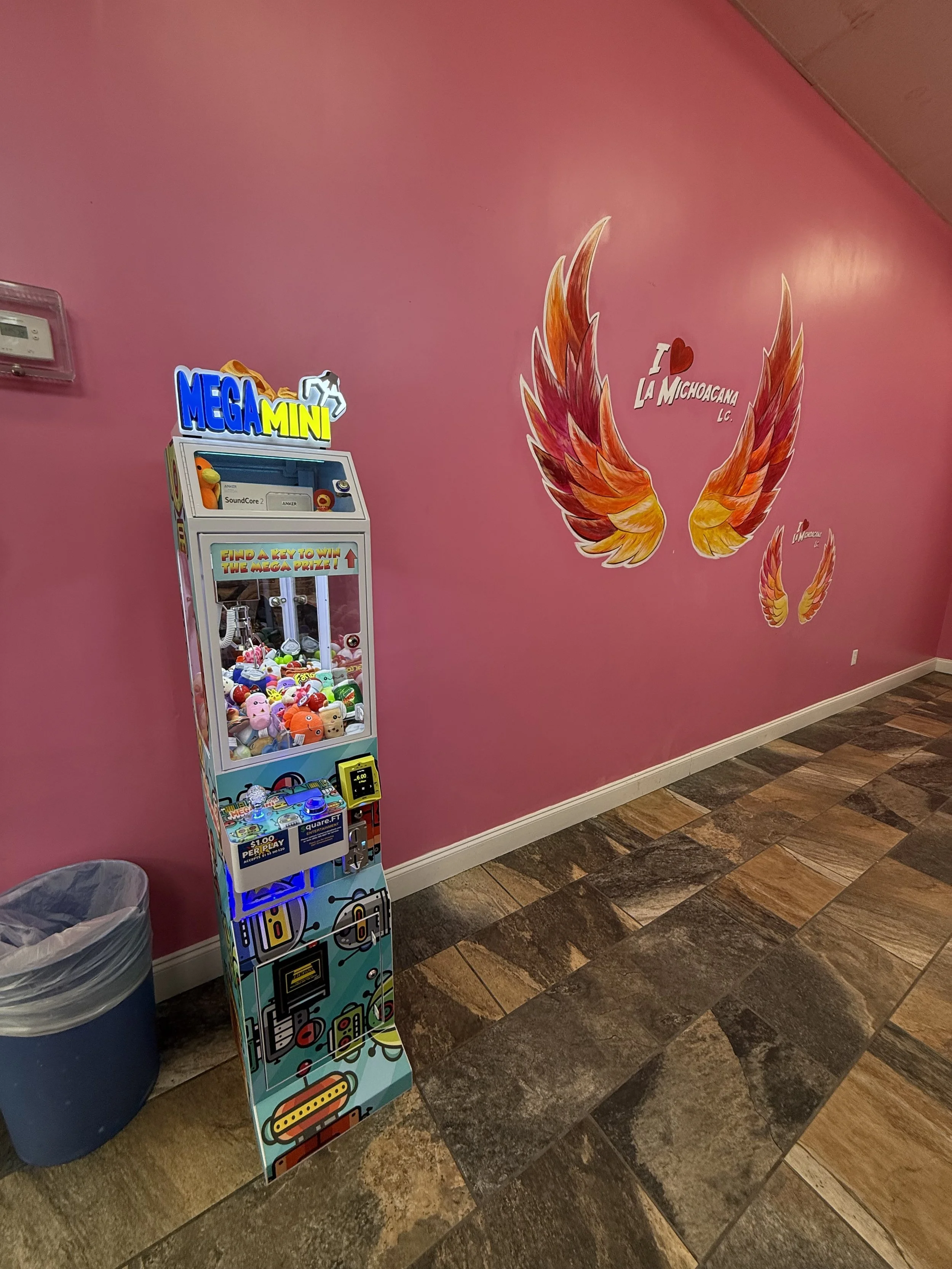 Pink wall in a room with a mural of wings and the text 'I love La Michoacana LC.' near the ceiling. A toy claw machine called 'Mega Mini' is on the floor next to a trash can. The floor has brown and beige tile patterns.