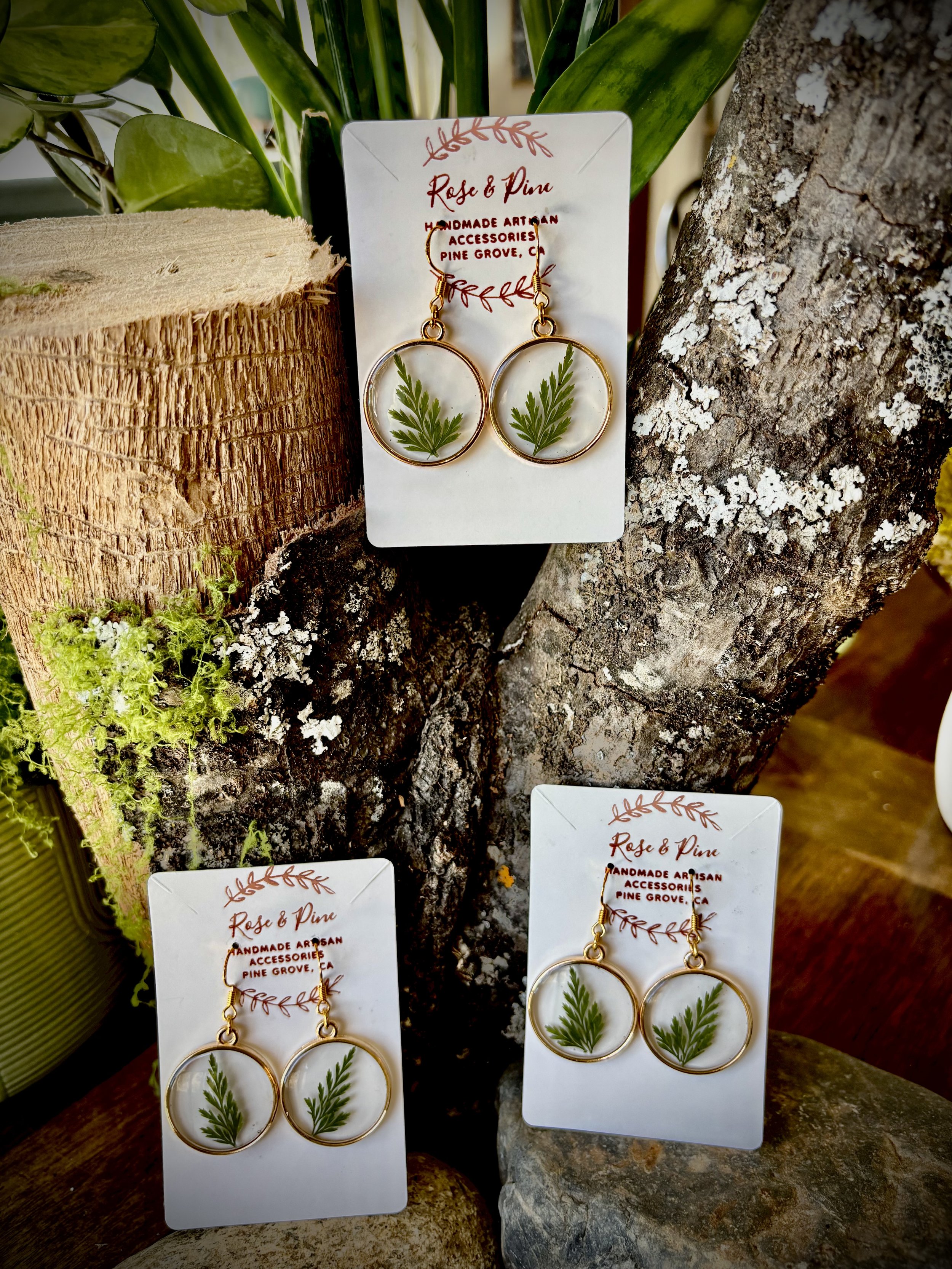 Three pairs of handmade earrings with green fern leaf designs, displayed on white cards labeled 'Rose & Pine,' attached to natural wood and tree bark background.