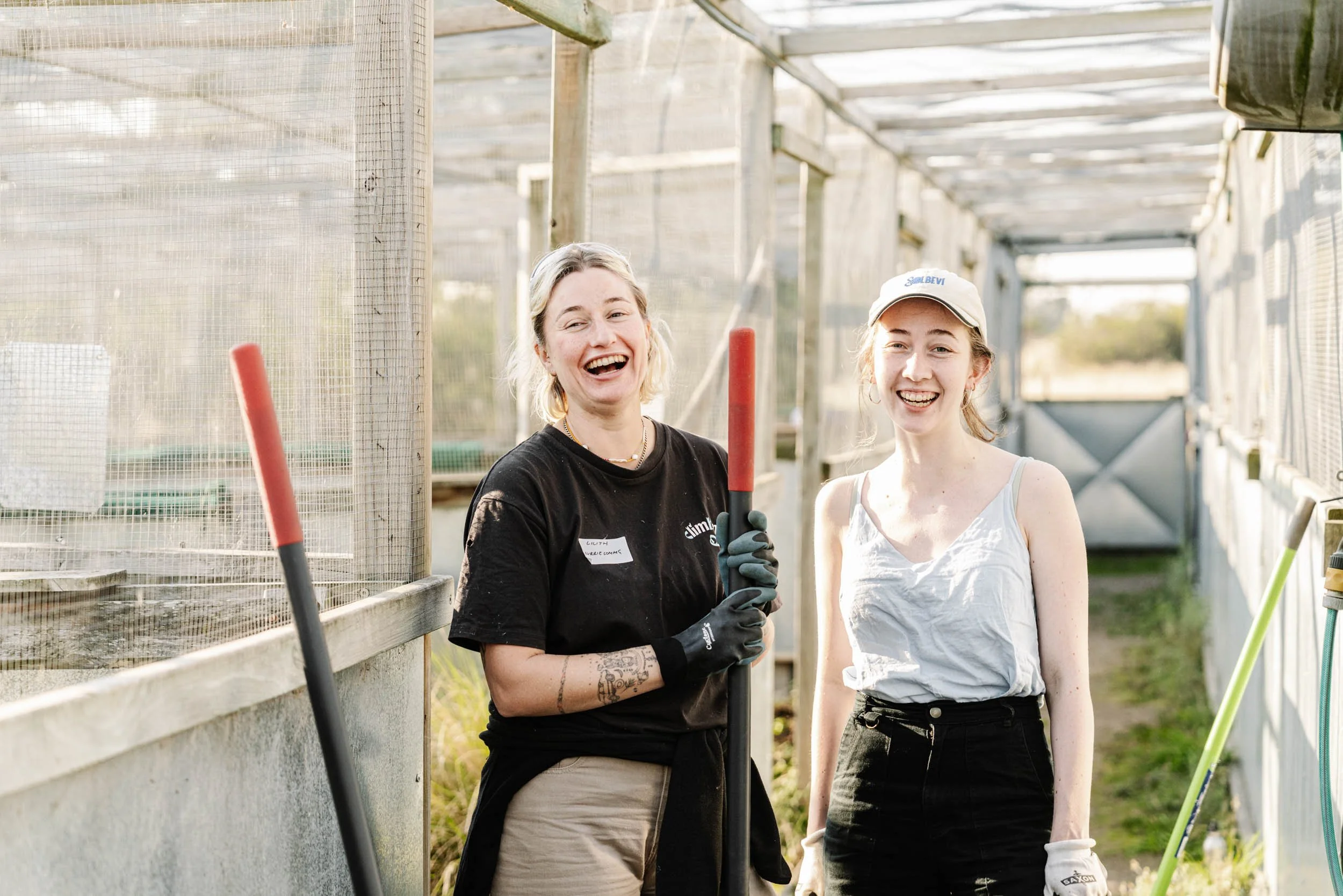 Two women smiling and holding gardening tools inside a greenhouse, wearing gloves and casual clothes.