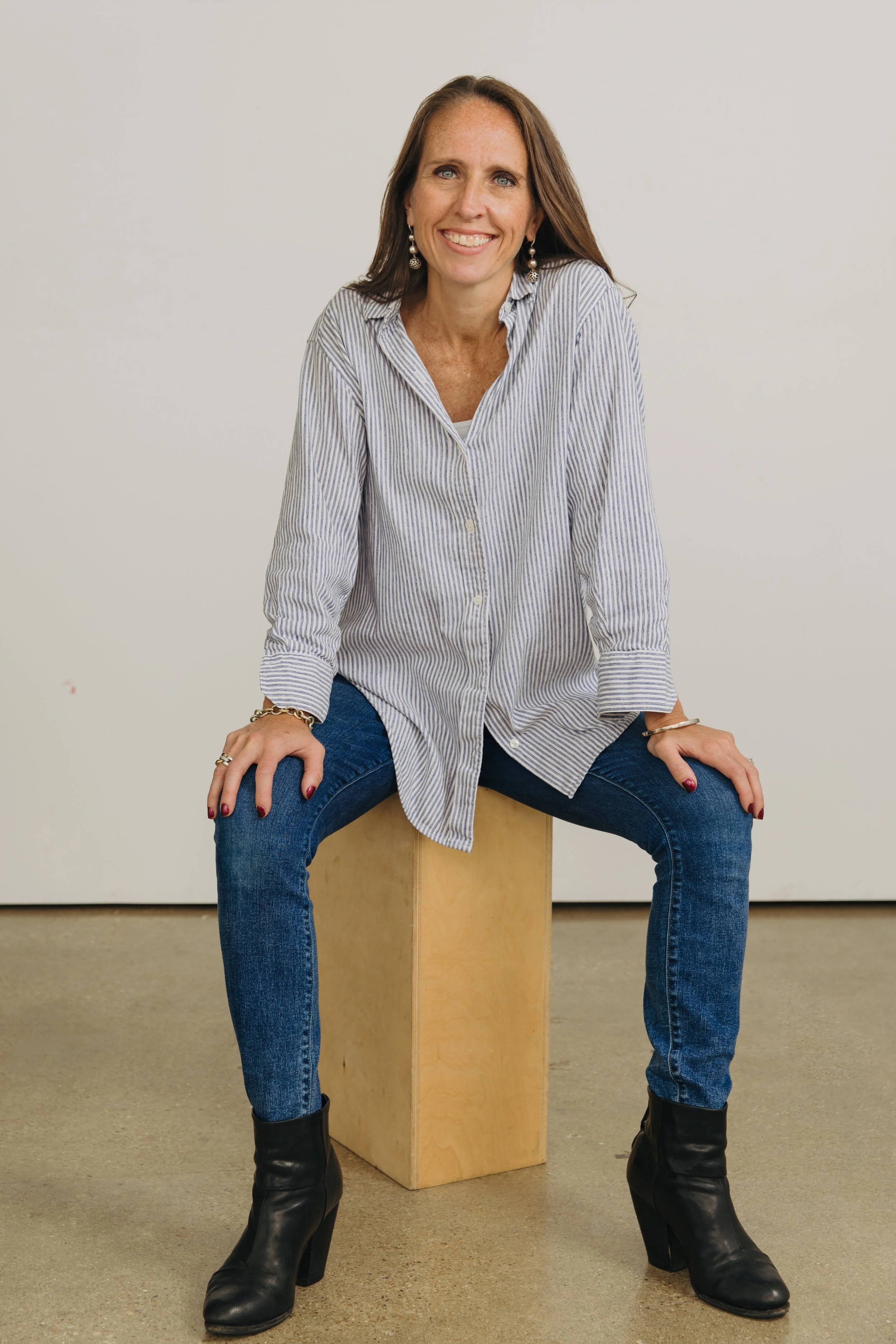 Woman with long brown hair, blue eyes, and earrings sitting on a wooden block in front of a white background, wearing a striped shirt, jeans, and black boots, smiling at the camera.