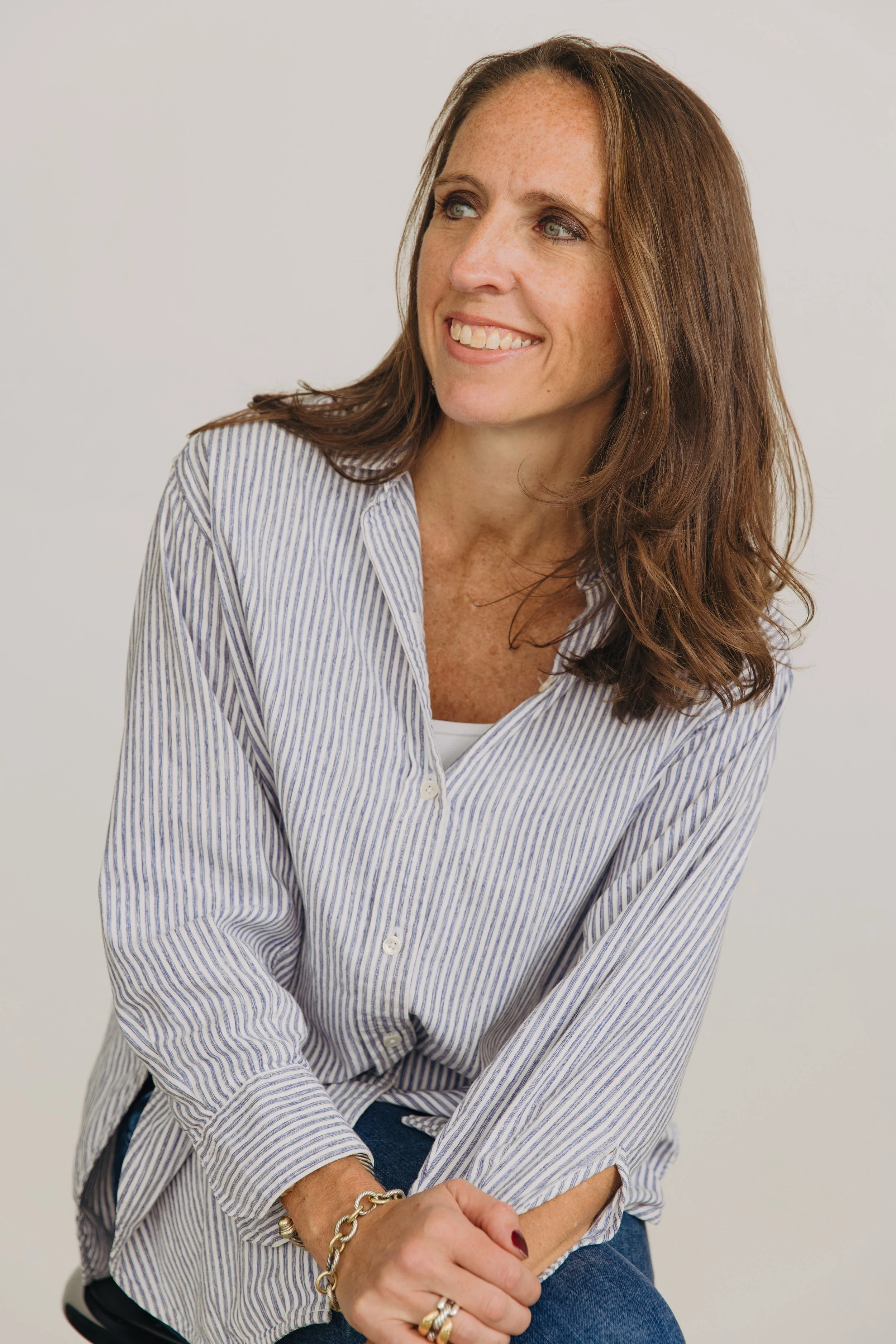 A woman with long brown hair and freckles smiling, wearing a striped button-up shirt and jeans, sitting against a plain light background.