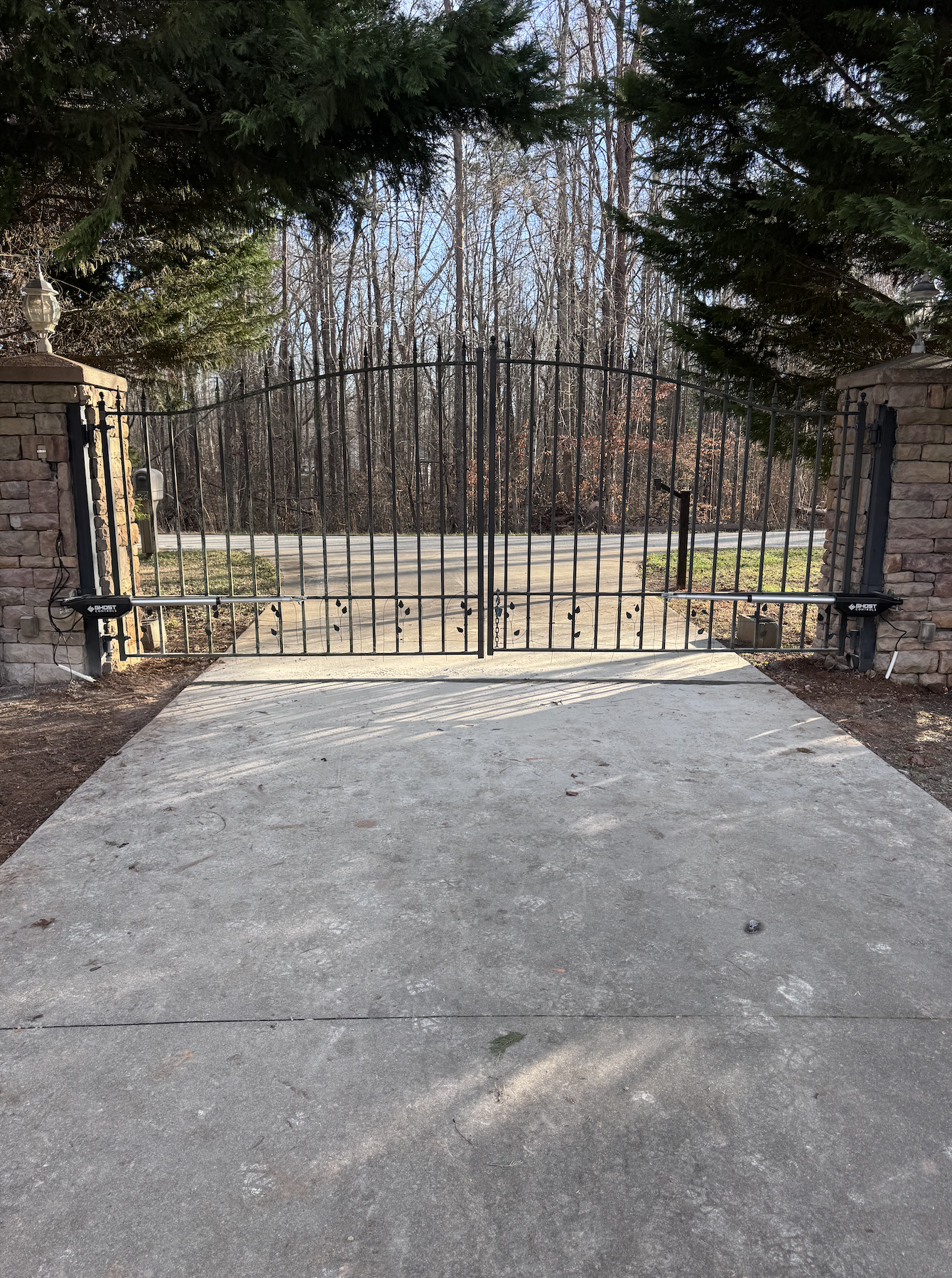 A closed metal gate set between two brick pillars at the entrance of a driveway, with a driveway in the foreground and a wooded area with bare trees in the background.