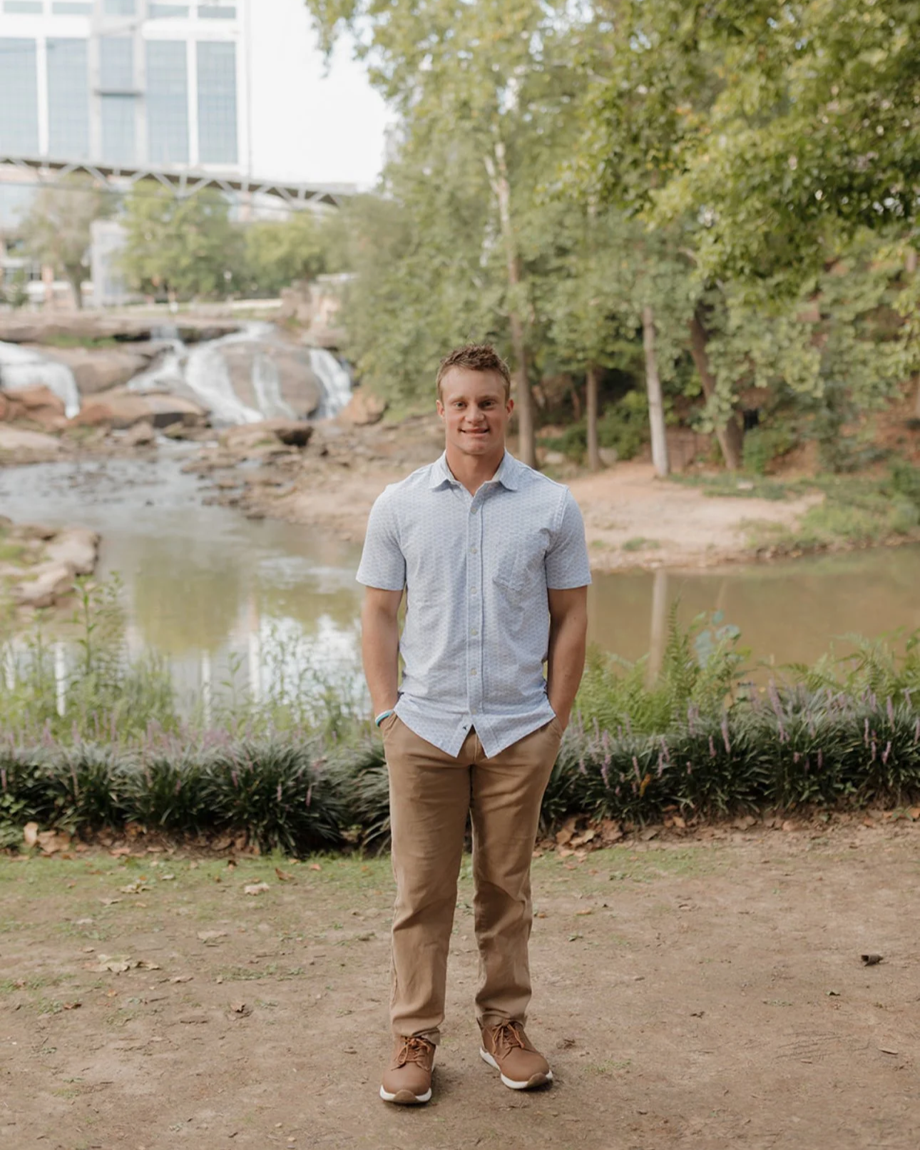 A young man standing outdoors near a river with rocks and trees, wearing a light blue short-sleeved shirt, tan pants, and brown shoes, smiling at the camera.