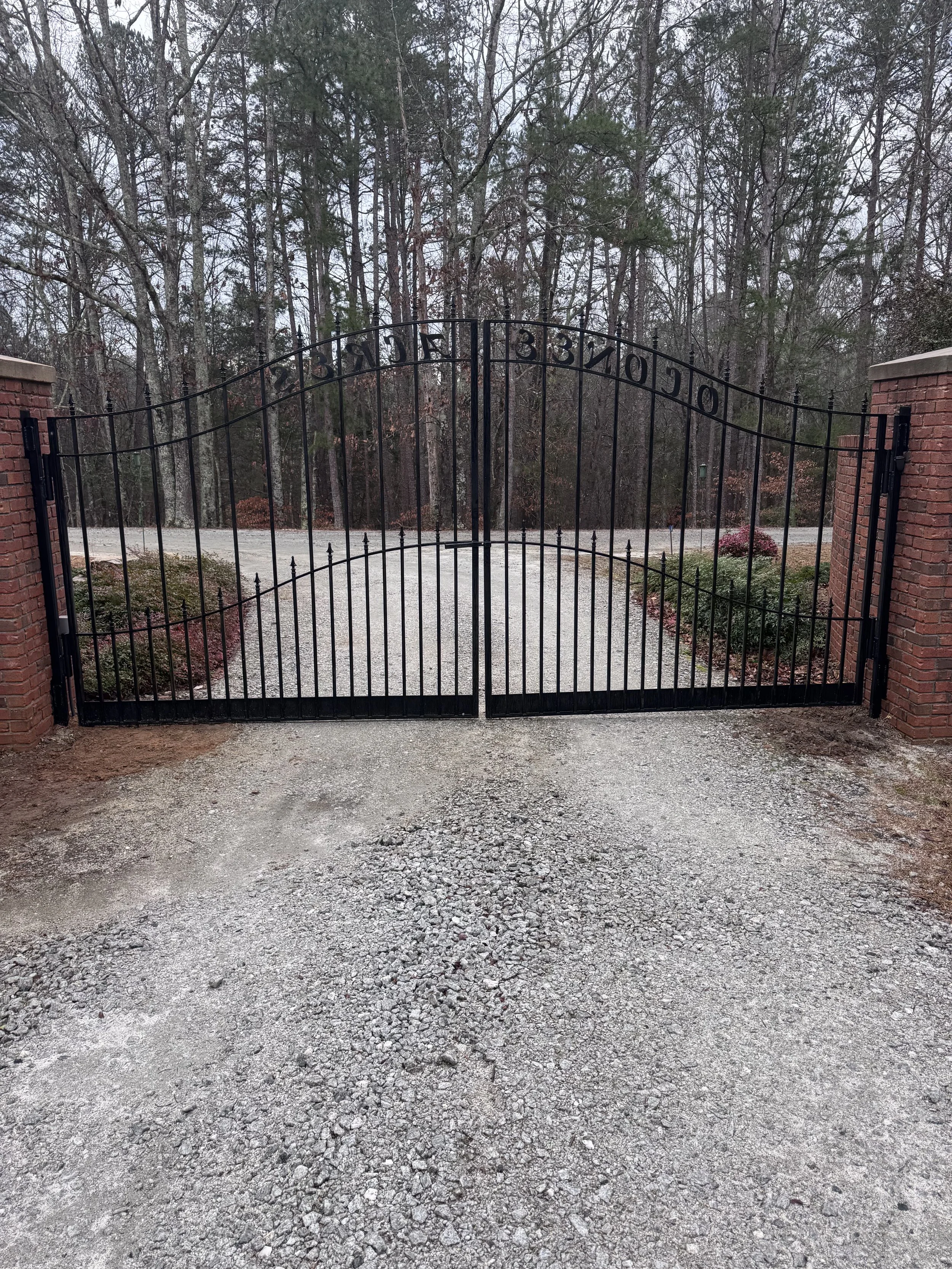 Black metal gate with house number 3310, flanked by brick pillars, leading to a gravel driveway into a wooded area with trees and shrubbery.