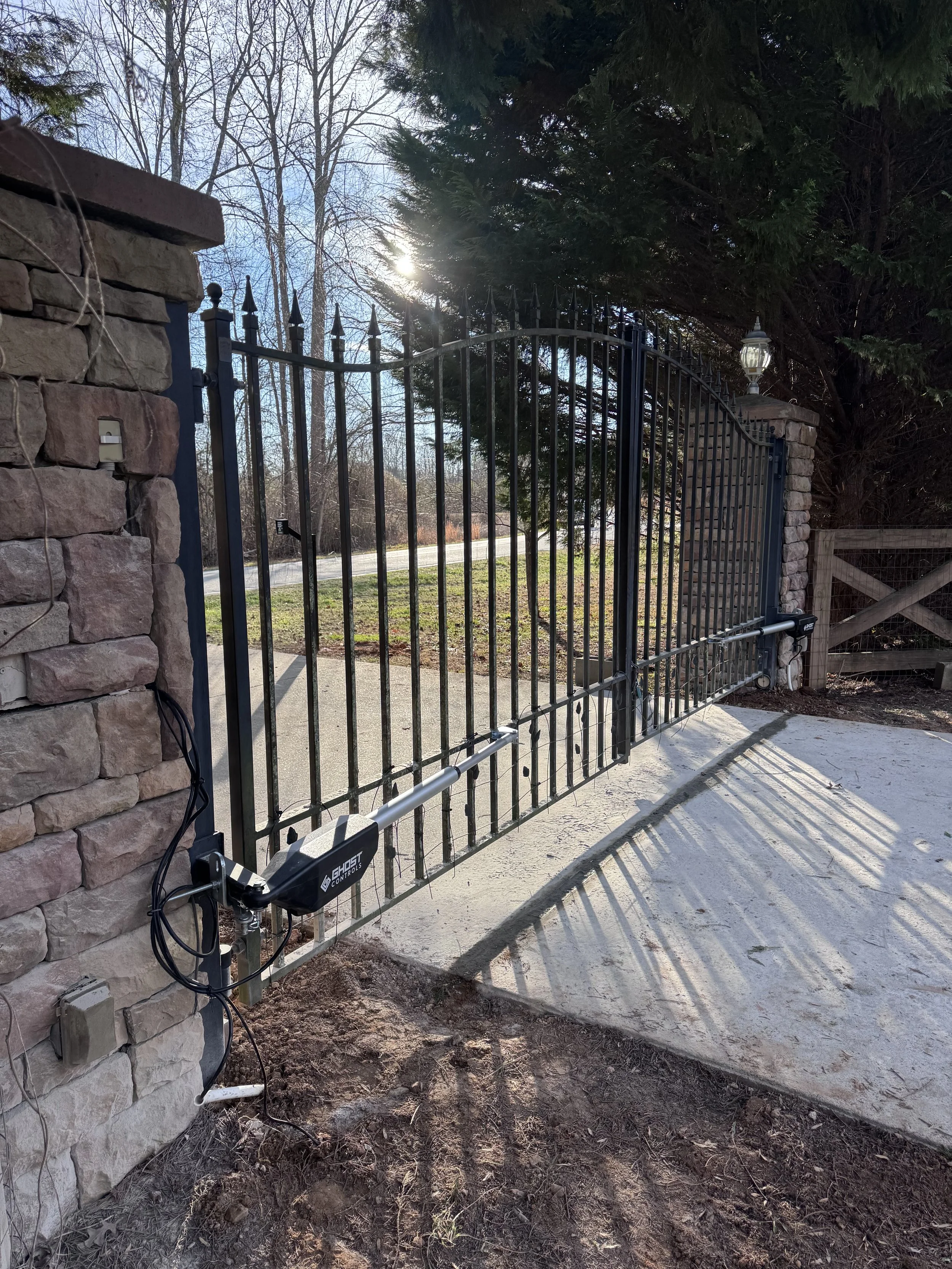 Automated driveway gate attached to stone pillars, with a motorized gate opener labeled Ghost Controls, partially open, casting shadows on concrete driveway, with trees and a fence in the background.