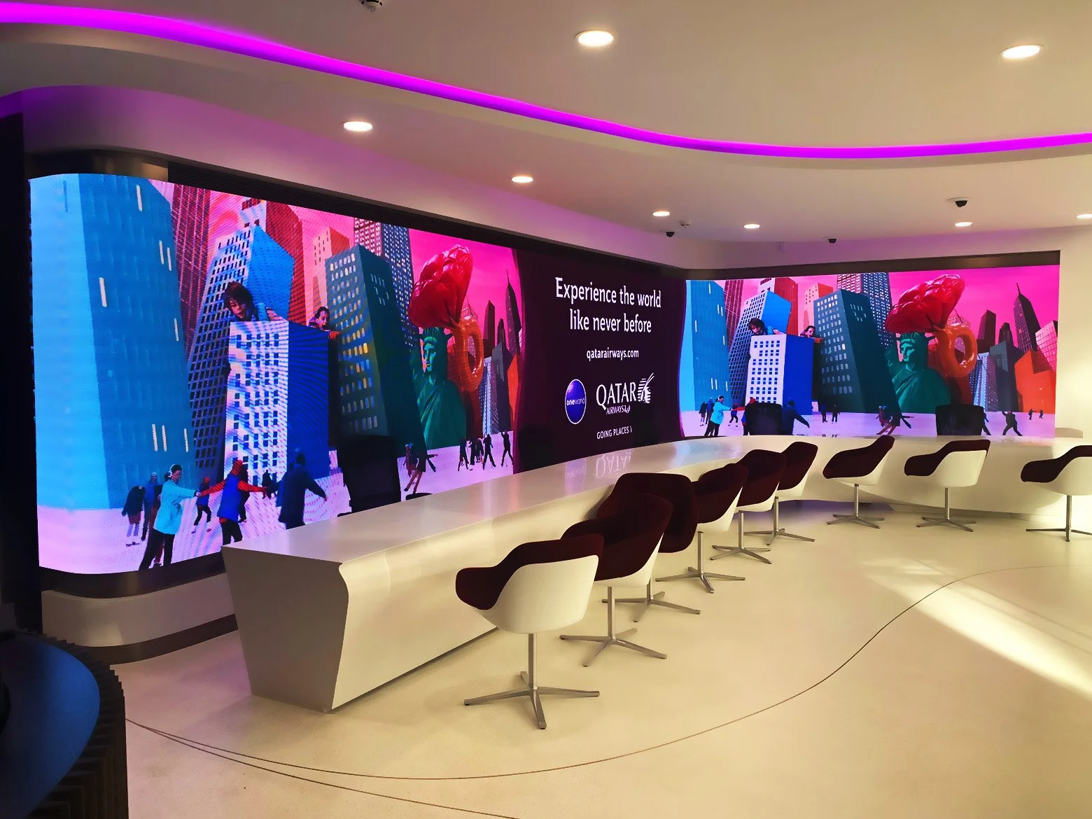 Large curved digital advertisement display in an airport lounge, showing vibrant images of a cityscape with tall buildings, a large Statue of Liberty figure, and people walking, with the text 'Experience the world like never before' and a Qatar Airways logo.