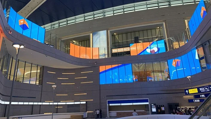 Interior of a modern multi-level building with glass walls and digital screens displaying blue and orange graphics.